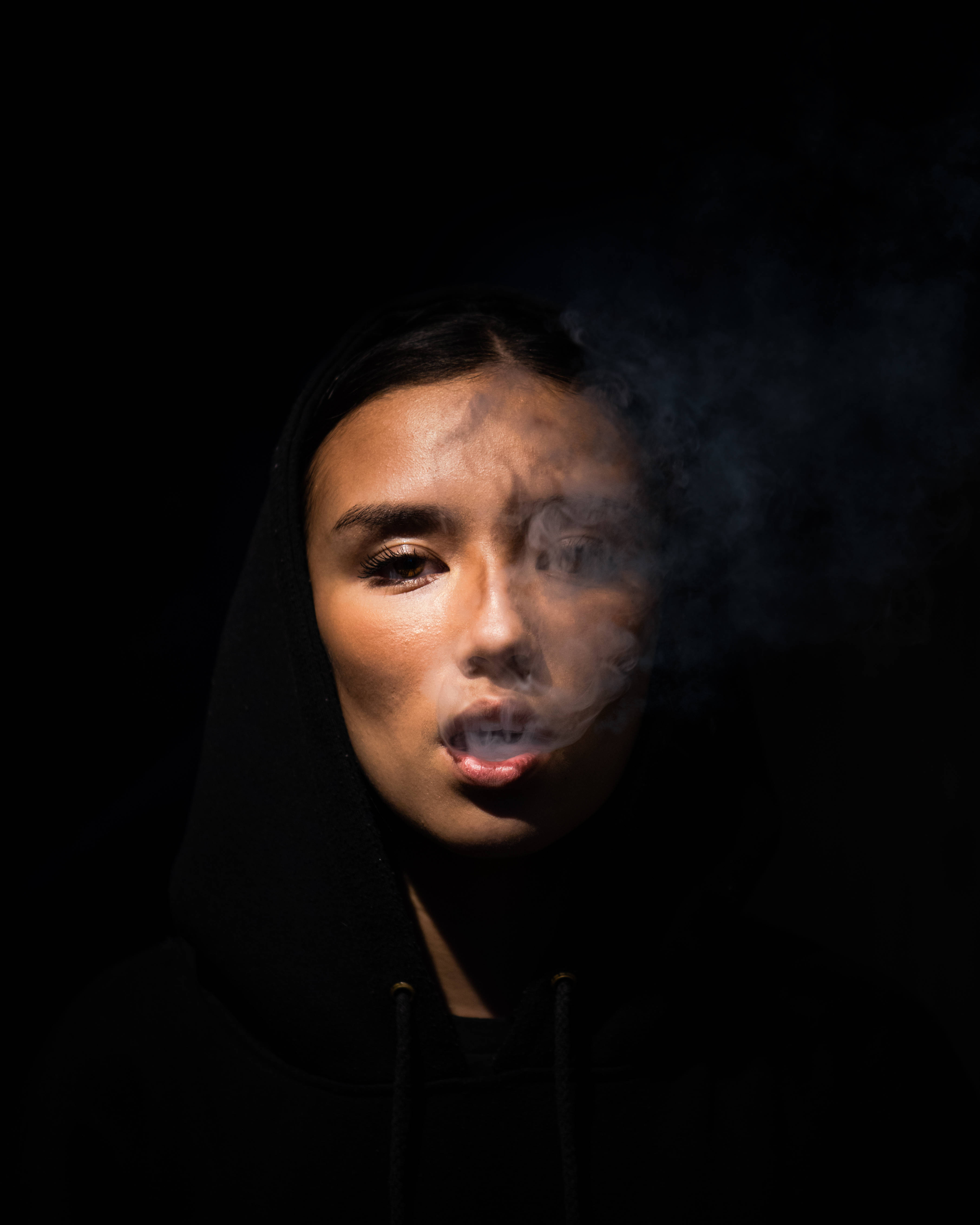 黑色美學 桌布 壁紙 — 一個穿著黑色連帽衫的人，在黑暗背景中戲劇性燈光下呼出煙霧。