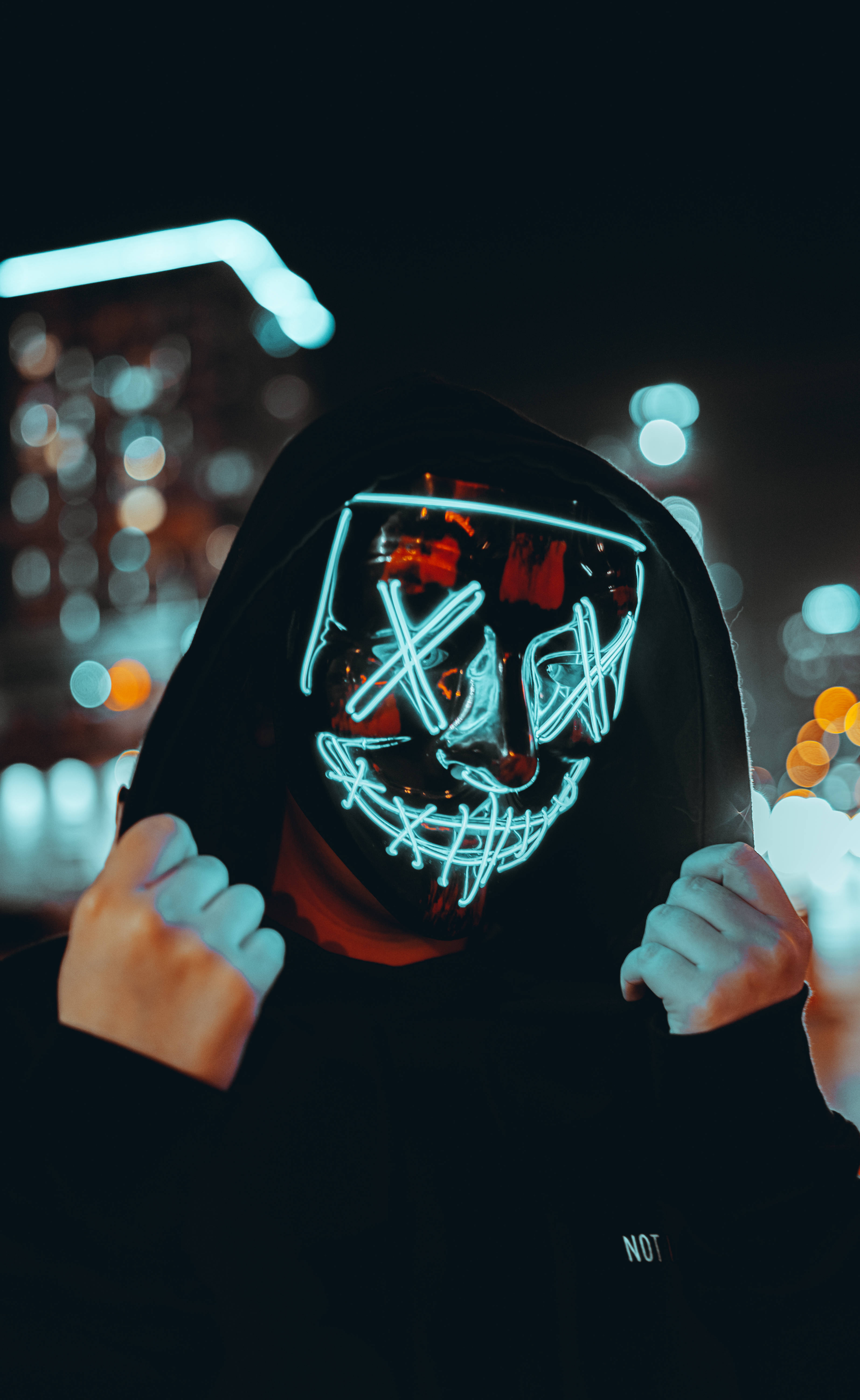 Purge 桌布 壁紙 — 一個穿著黑色連帽衫的人，戴著發光藍色LED面具，露出邪惡的笑容，背景是模糊的城市夜景。
