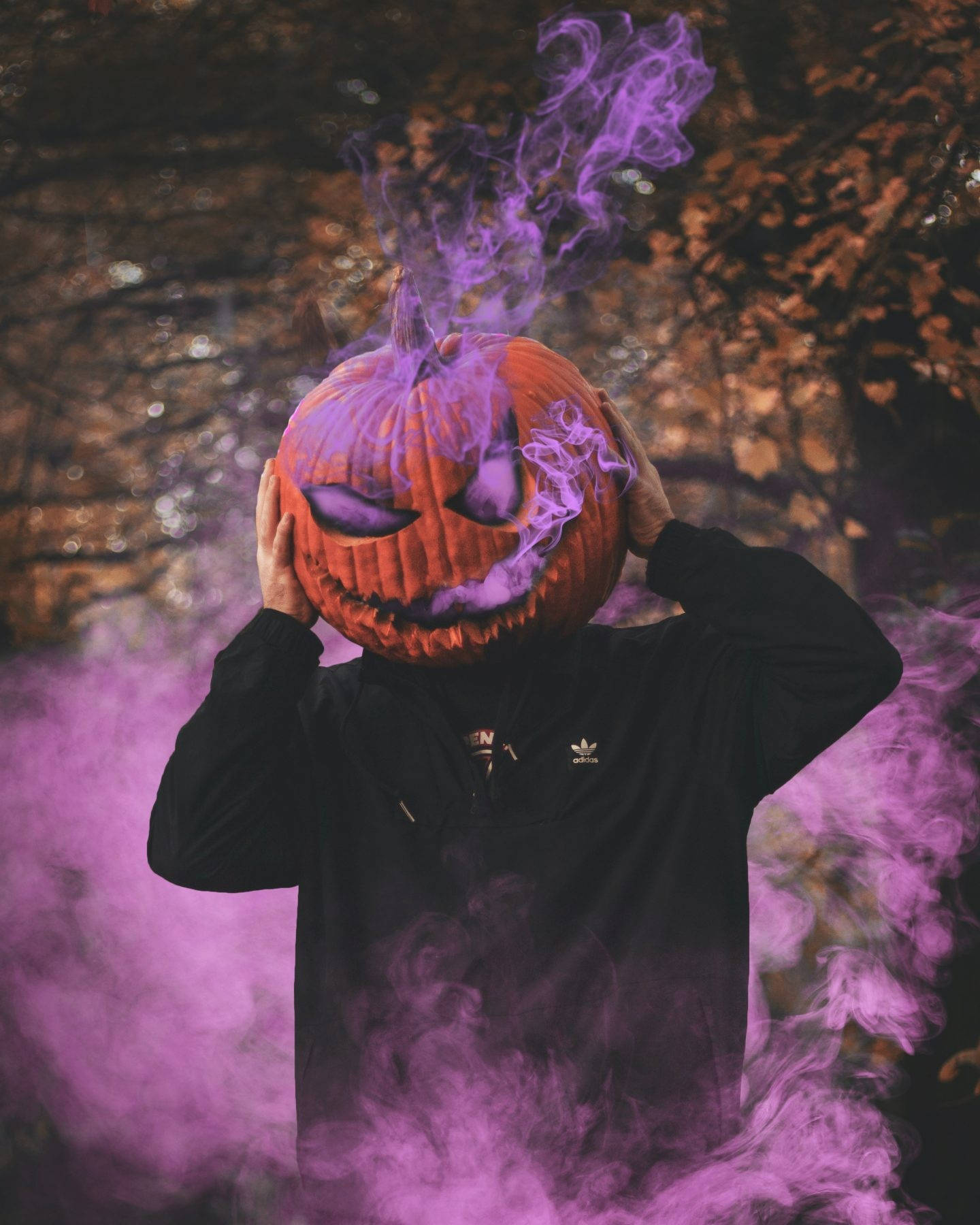 Spooky Aesthetic 桌布 壁紙 — 一個穿著黑色連帽衫的人手持一個雕刻的南瓜頭，散發出旋轉的紫色煙霧，背景是模糊的秋日森林。