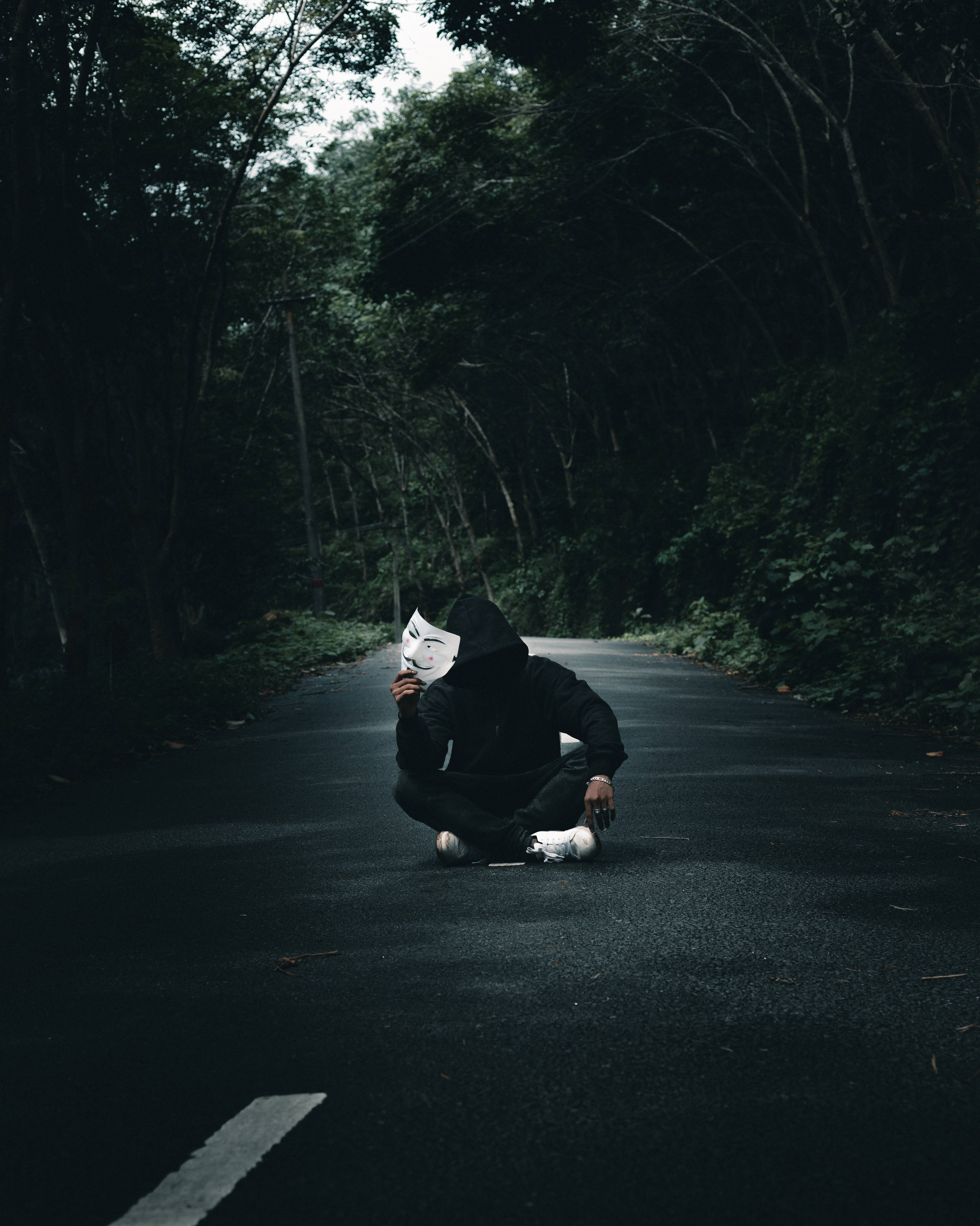 匿名 桌布 壁紙 — 一個穿著黑色連帽衫的人獨自坐在黑暗的森林路上，手持白色Anonymous面具。