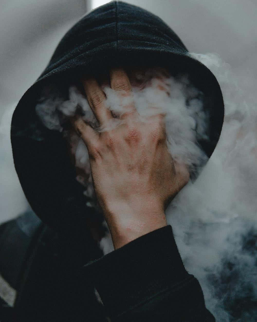 Instagram Profile 桌布 壁紙 — 一個穿著黑色連帽衫的人用手遮住臉，濃厚的白色蒸氣在他們周圍旋轉，營造出陰鬱的特寫鏡頭。