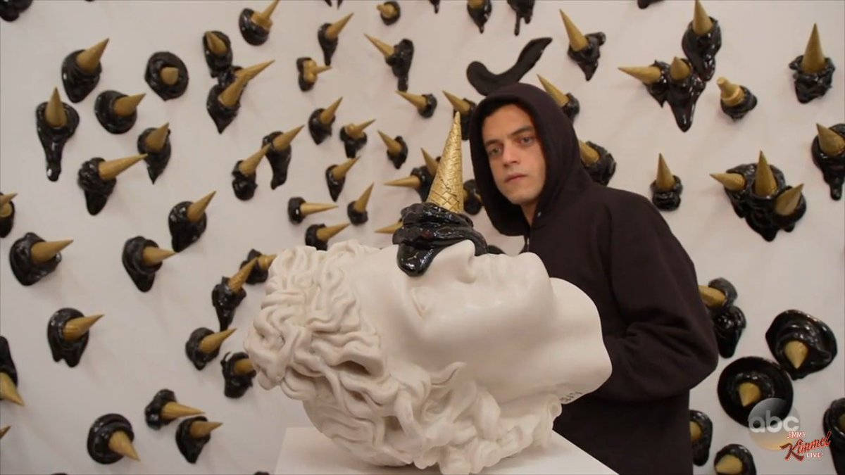 Rami Malek 桌布 壁紙 — 一個穿著黑色連帽衫的人站在一個白色大理石半身像旁邊，身後牆上有一個金色角和黑色黏糊糊的角。