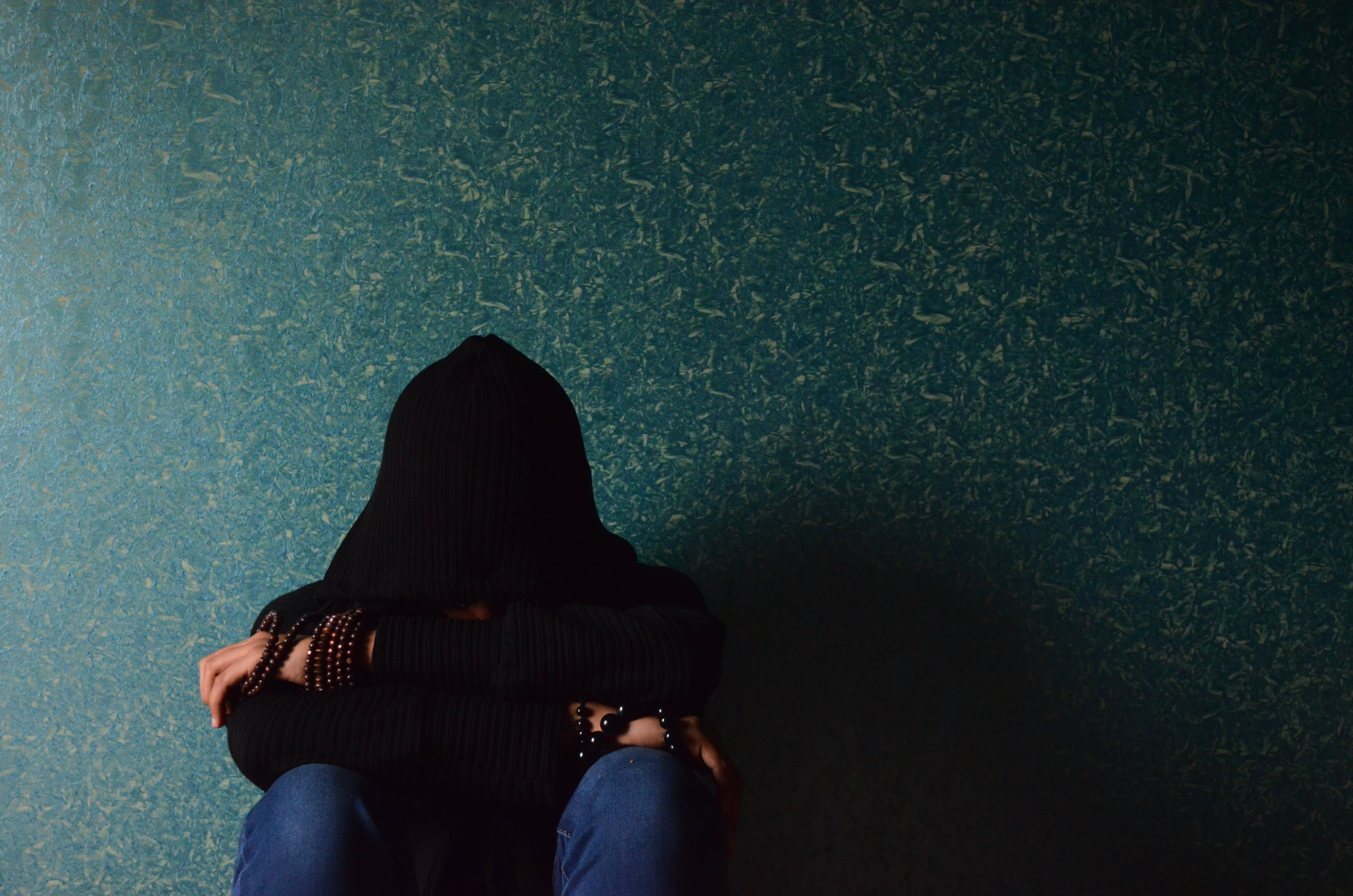 Depression Aesthetic 桌布 壁紙 — 一個穿著黑色連帽衫的人蜷縮著坐在有紋理的青綠色牆壁旁，雙臂環抱膝蓋，顯得孤獨。