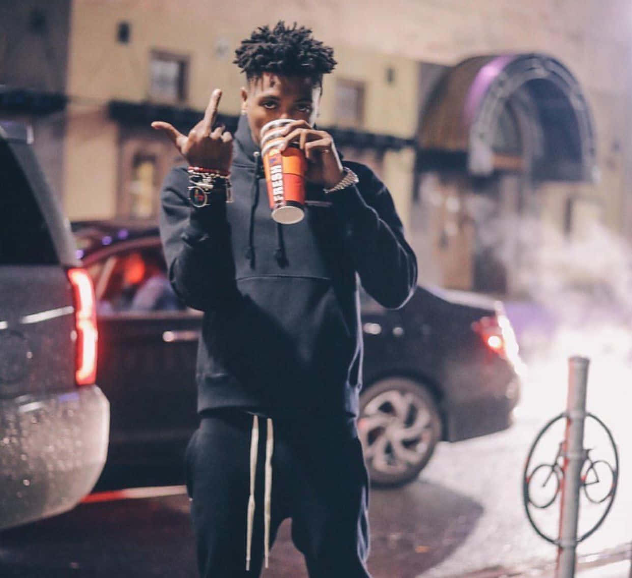 Youngboy 桌布 壁紙 — 一個穿著黑色連帽衫的年輕男子在雨夜的城市街道上從紅色杯子中啜飲並做出手勢。
