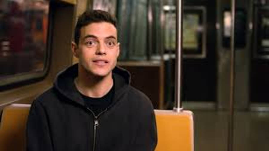Rami Malek 桌布 壁紙 — 一個穿著黑色連帽衫的年輕男子靜靜地坐在地鐵座位上，面無表情地直視鏡頭。