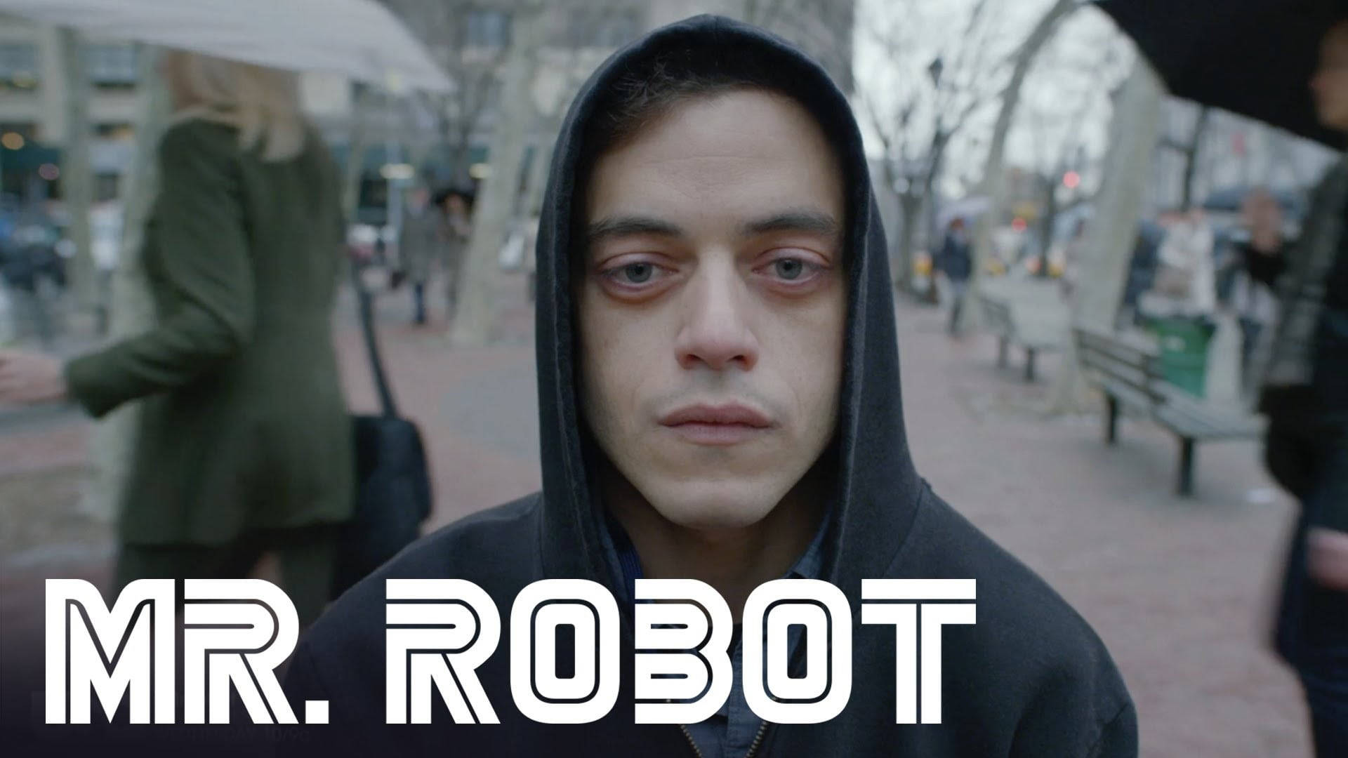 Mr Robot 桌布 壁紙 — 一個穿著黑色連帽衫的男人在雨天的城市人行道上，背景是模糊的行人和光禿禿的樹木，他正強烈地凝視著鏡頭。