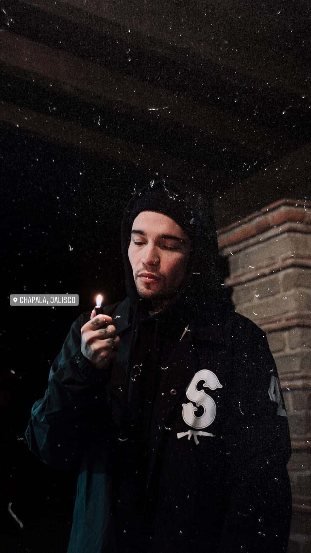 Hoodie 桌布 壁紙 — 一個穿著黑色連帽衫的男人在雪夜背景下，手持點燃的打火機，背景是磚砌建築。