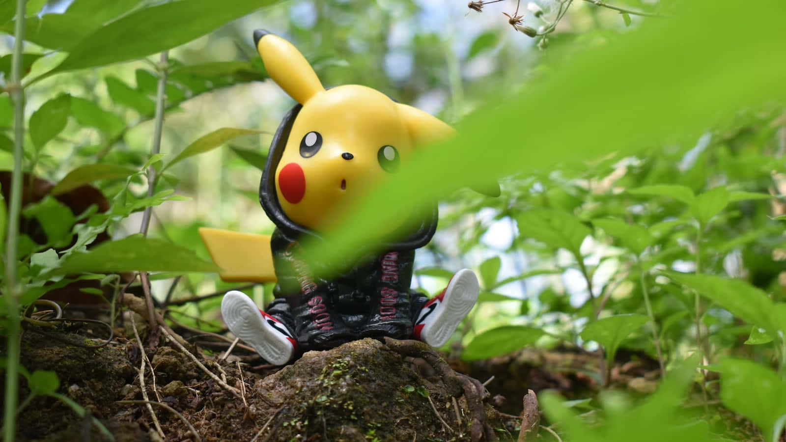 Pokemon Toys 桌布 壁紙 — 一個穿著黑色連帽衫的黃色皮卡丘公仔坐在長滿青苔的石頭上，周圍是茂密的綠色植物。