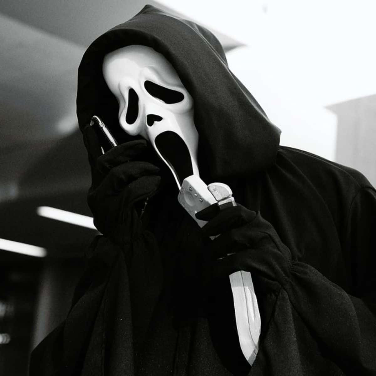 鬼臉殺手 桌布 壁紙 — 一個穿著黑色連帽長袍、戴著標誌性白色尖叫面具的人，在昏暗的室內空間中一手持刀，一手緊握電話。