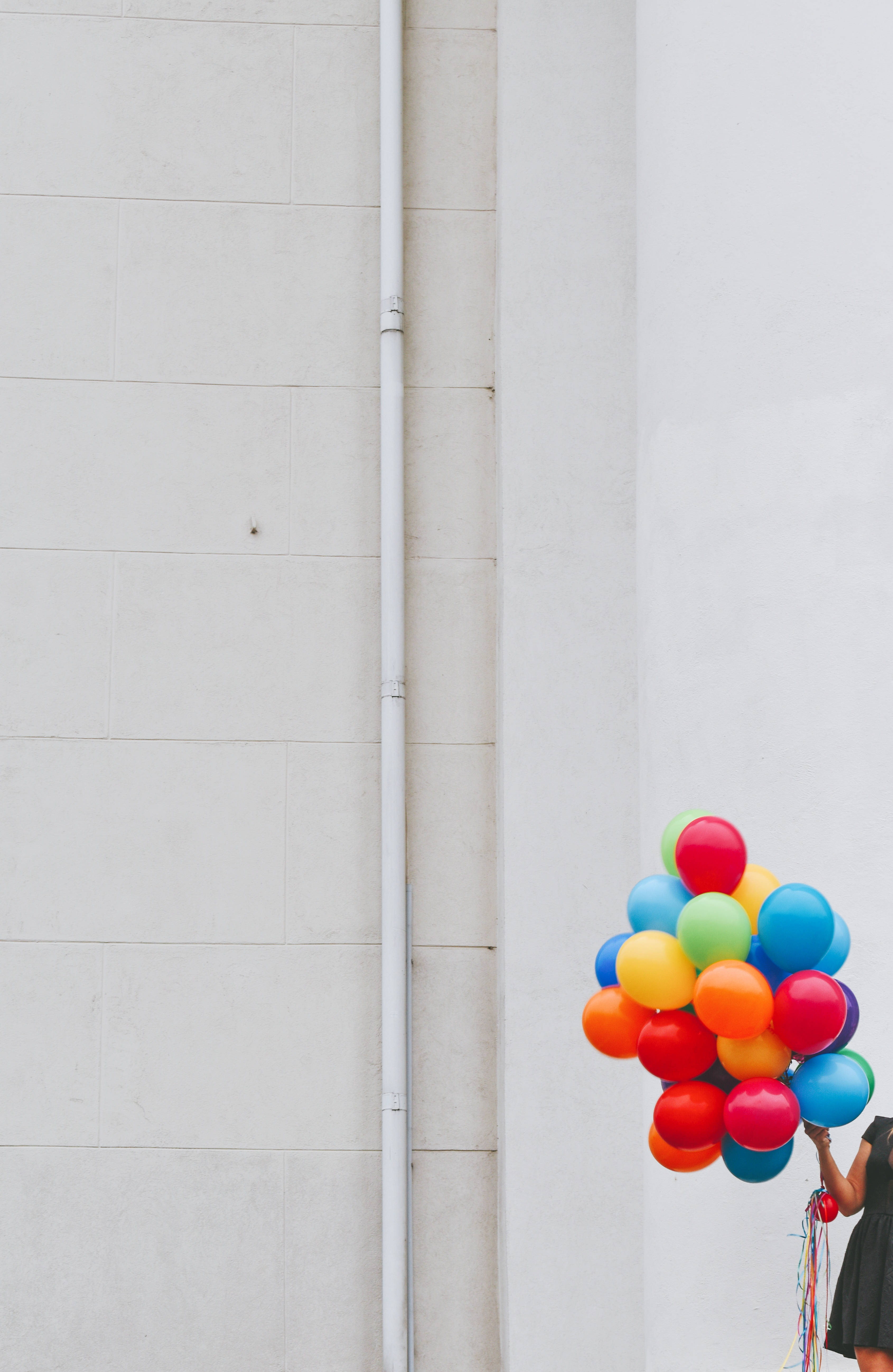 Balloon 桌布 壁紙 — 一個穿著黑色連衣裙的人，在純白色的城市牆壁前，拿著一束色彩繽紛的氣球。