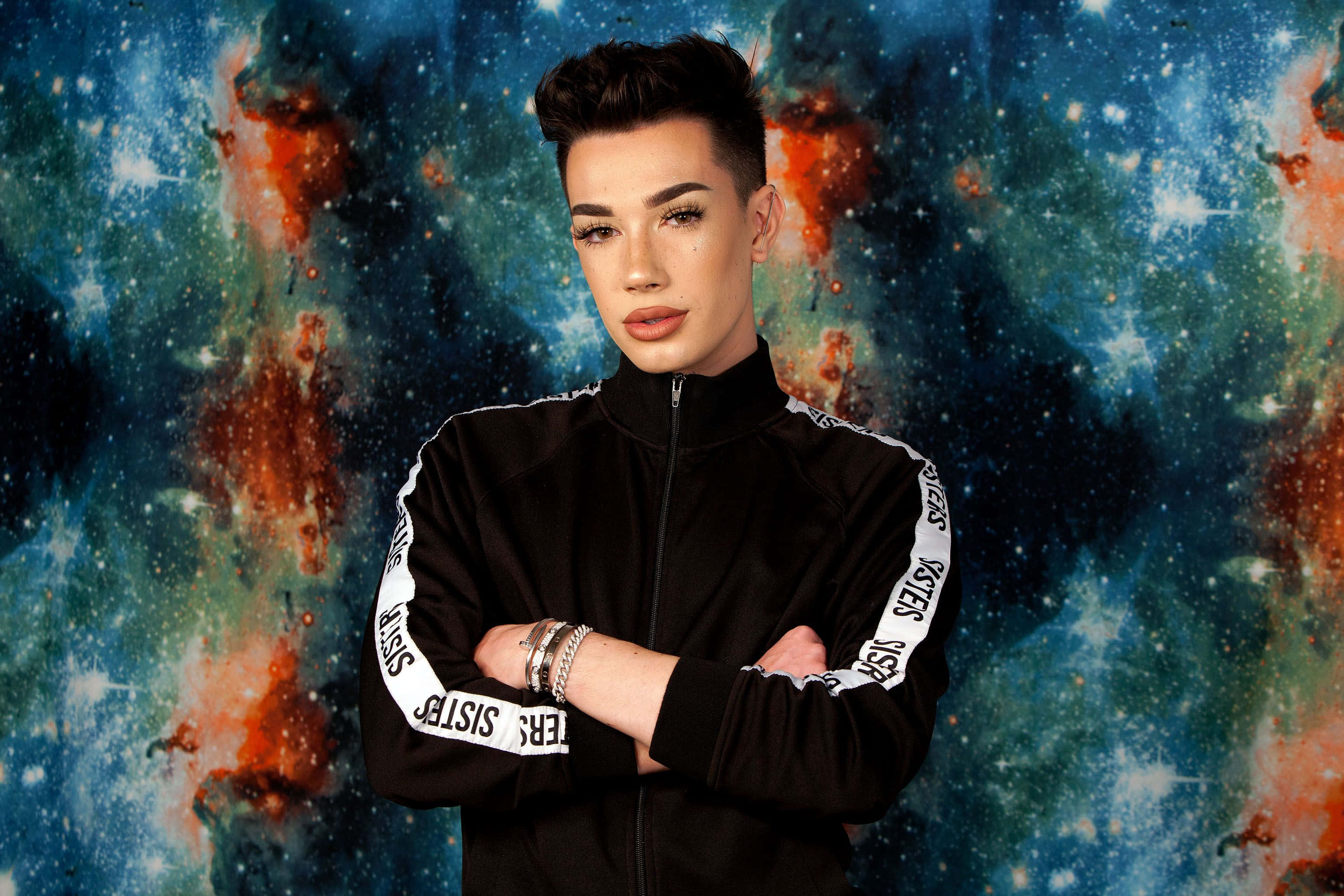 James Charles 桌布 壁紙 — 一個穿著黑色運動外套的人自信地站在一個充滿活力的宇宙星雲背景前，背景中有旋轉的藍色和橙色。