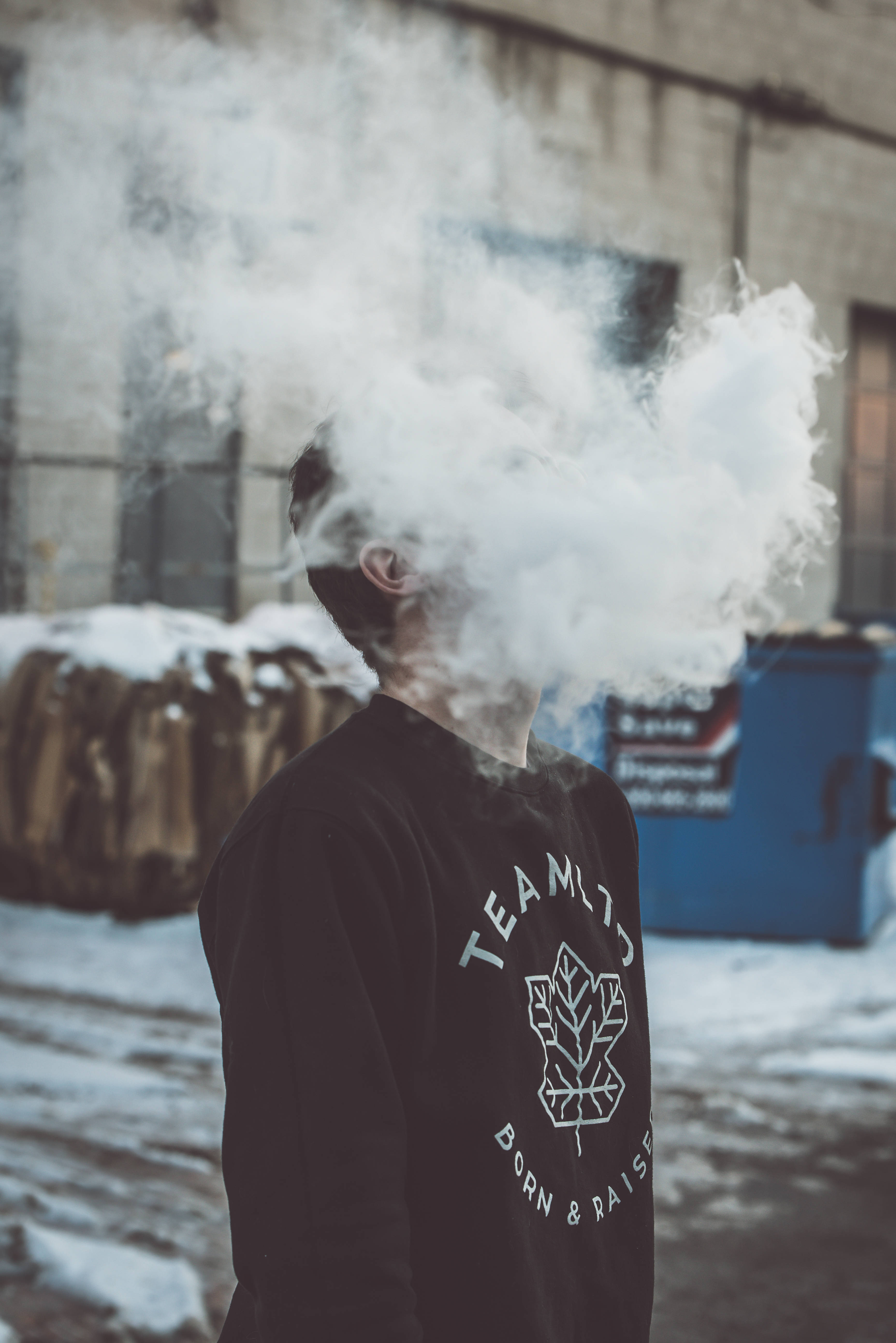 抽菸 桌布 壁紙 — 一個穿著黑色運動衫的人呼出一團濃密的蒸氣，遮蓋了他們的臉，背景是雪白的城市。