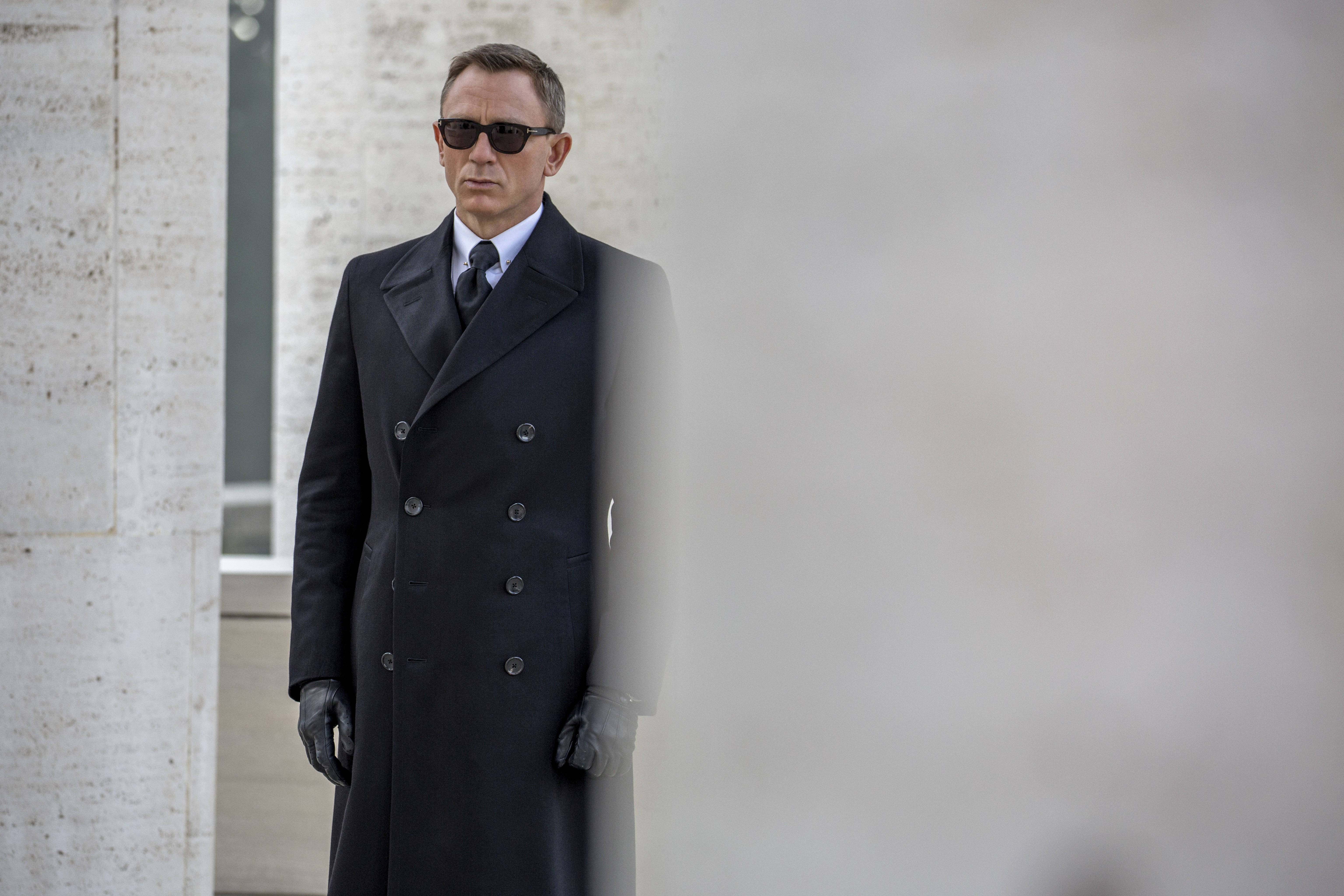 Daniel Craig 桌布 壁紙 — 一個穿著黑色雙排扣大衣和太陽眼鏡的男人，莊嚴地站在有紋理的混凝土牆前。