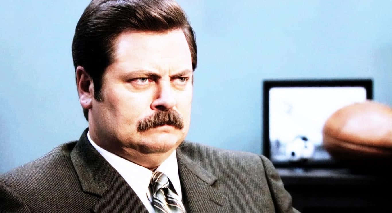 nick offerman 桌布 壁紙 — 一個穿西裝留鬍子的男人在辦公室裡，背景有足球和顯示器，他凝視著，神情專注。