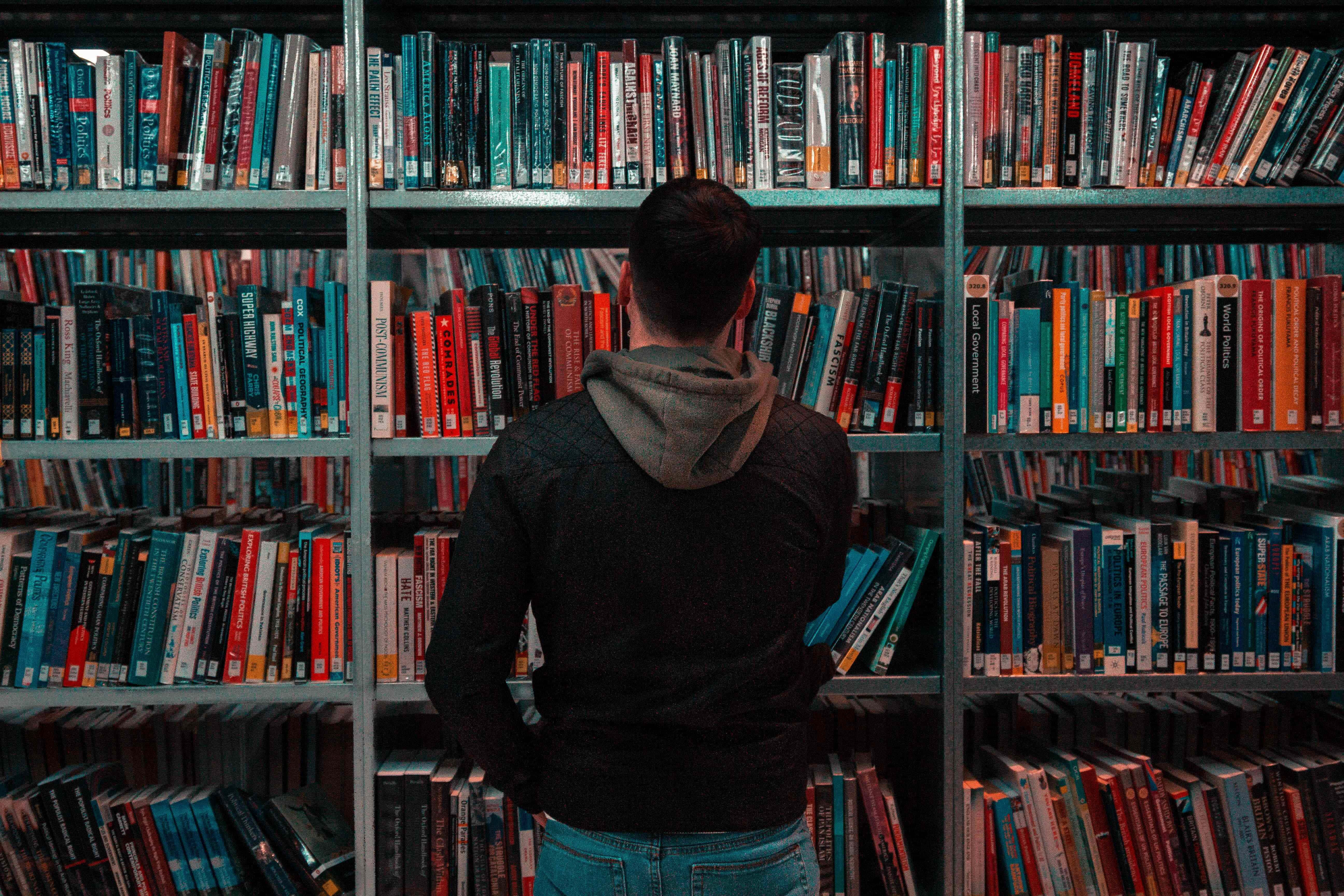 Learning 桌布 壁紙 — 一個穿連帽衫的人站在光線昏暗的圖書館裡，面對金屬書架上色彩繽紛的書本。