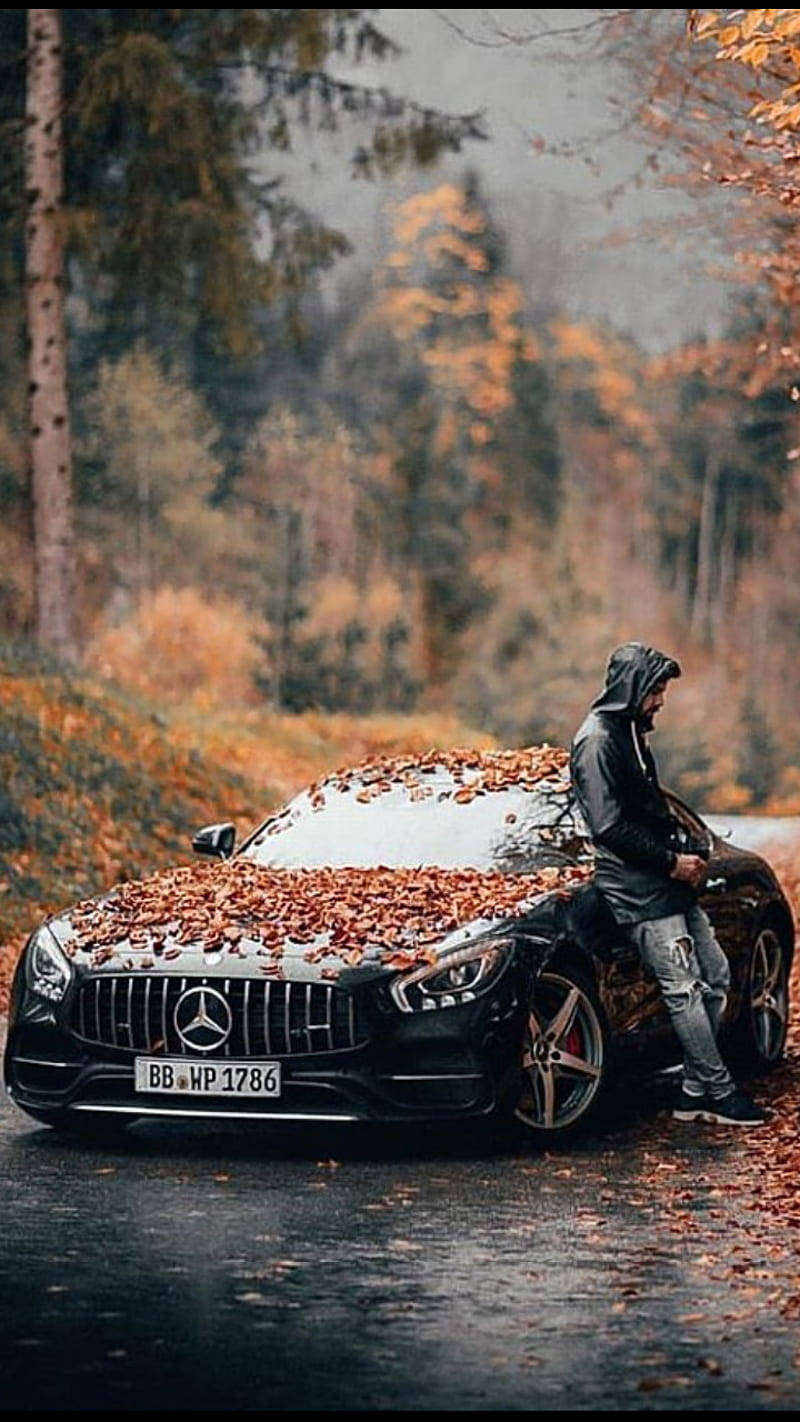 Mercedes 桌布 壁紙 — 一個穿連帽衫的人靠在一輛黑色賓士AMG上，車上鋪滿了秋天的落葉，在潮濕的森林道路上。