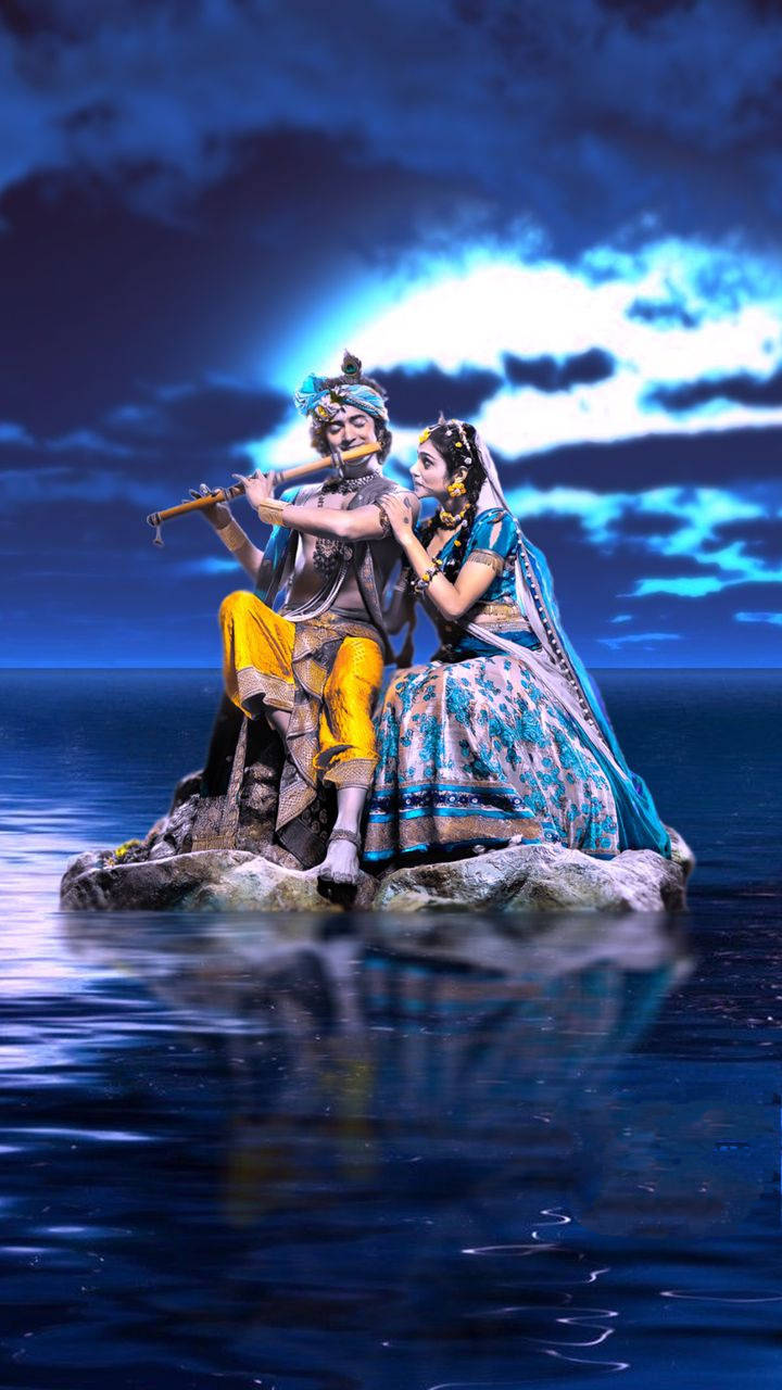 krishna iPhone 桌布 壁紙 — 一個穿黃色褲子的男人在吹笛子，而一個穿藍色紗麗的女人在月光下的深藍色水中岩石上聆聽。