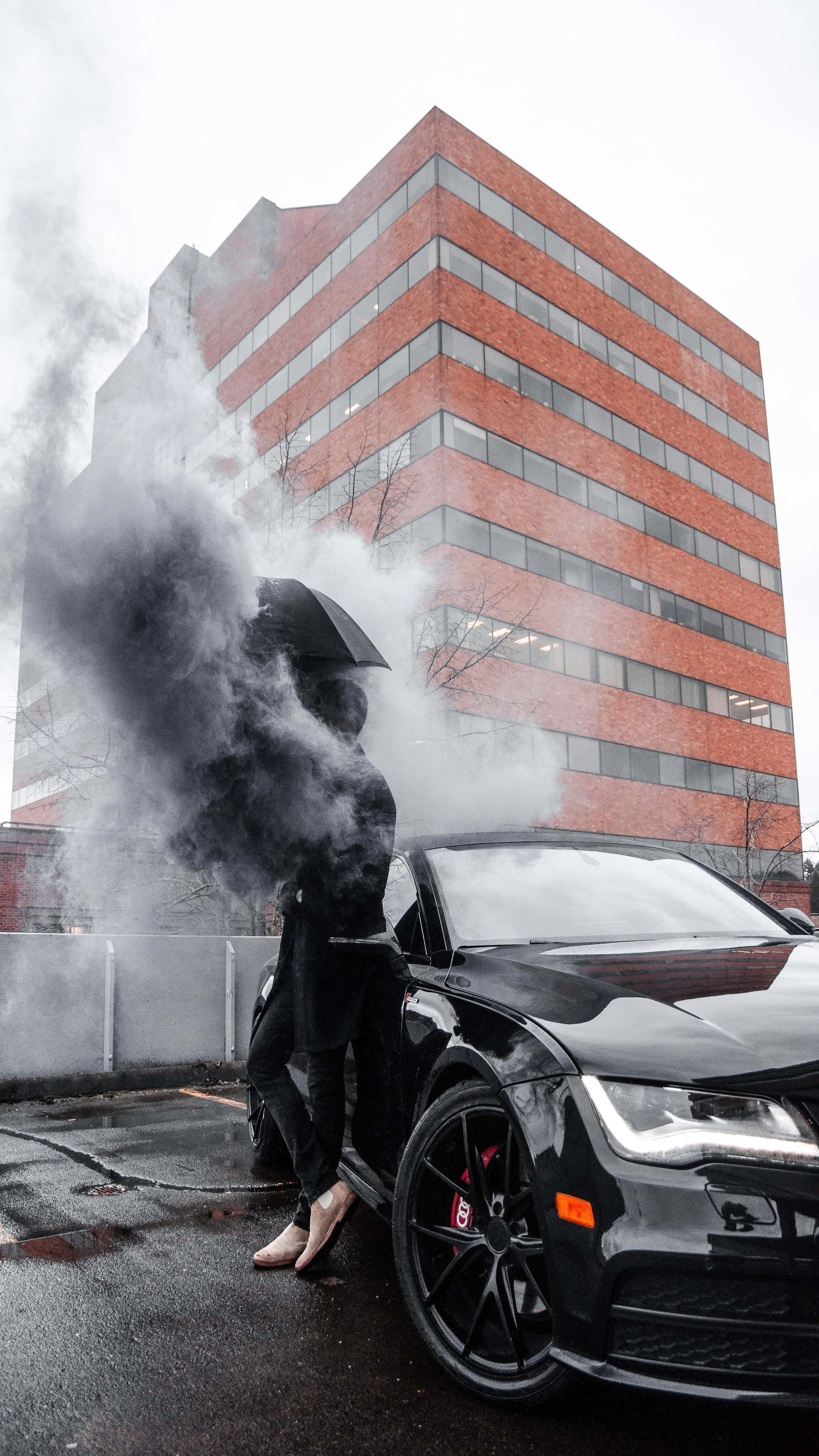 4K汽車 桌布 壁紙 — 一個穿黑色衣服的人在雨天，站在一個煙霧繚繞的雨傘下，在一棟紅磚建築前，站在一輛時尚黑色跑車旁邊。