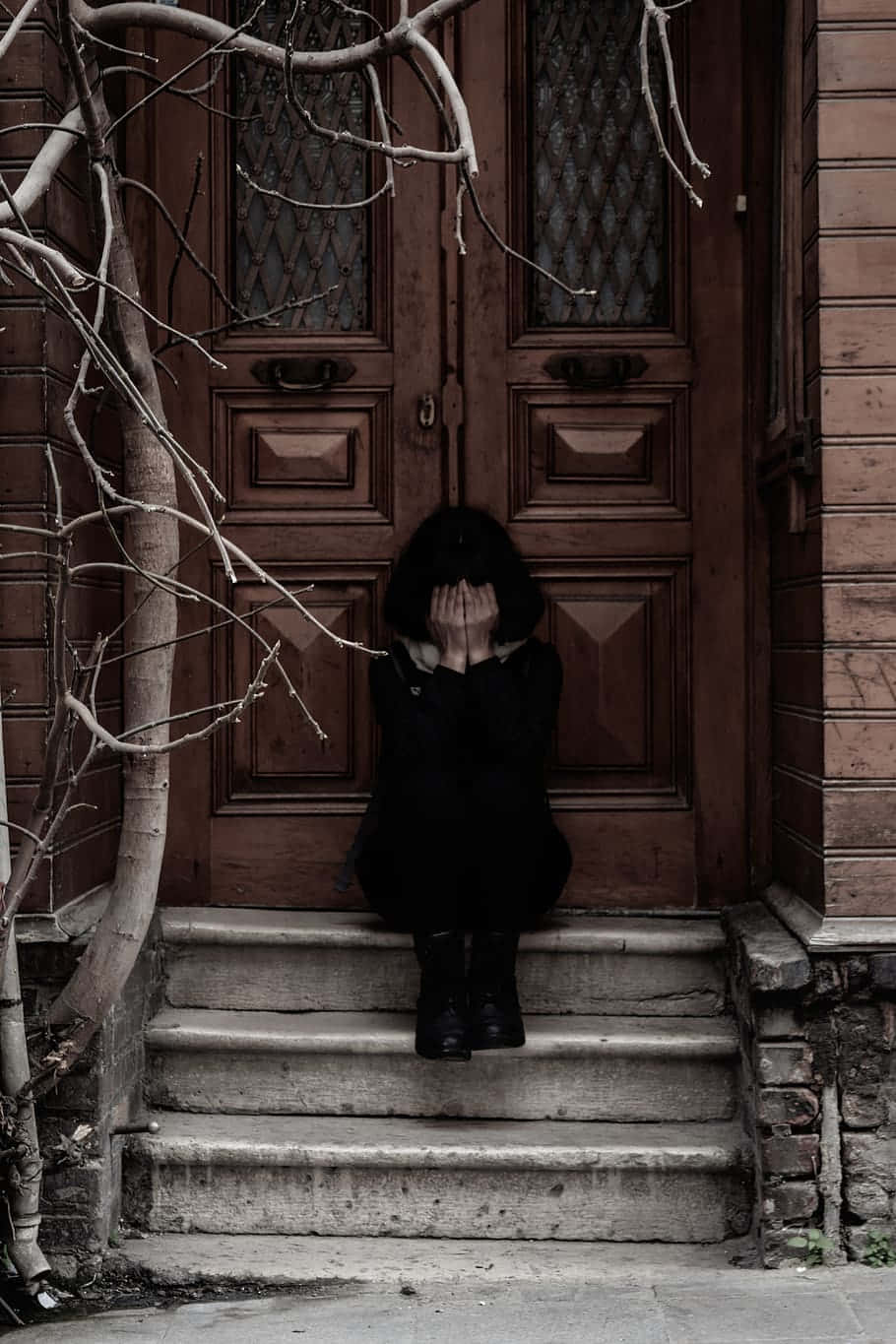 sad crying 桌布 壁紙 — 一個穿黑色衣服的人坐在石階上，在一個被冬日光禿樹枝環繞的風化木門前掩面。