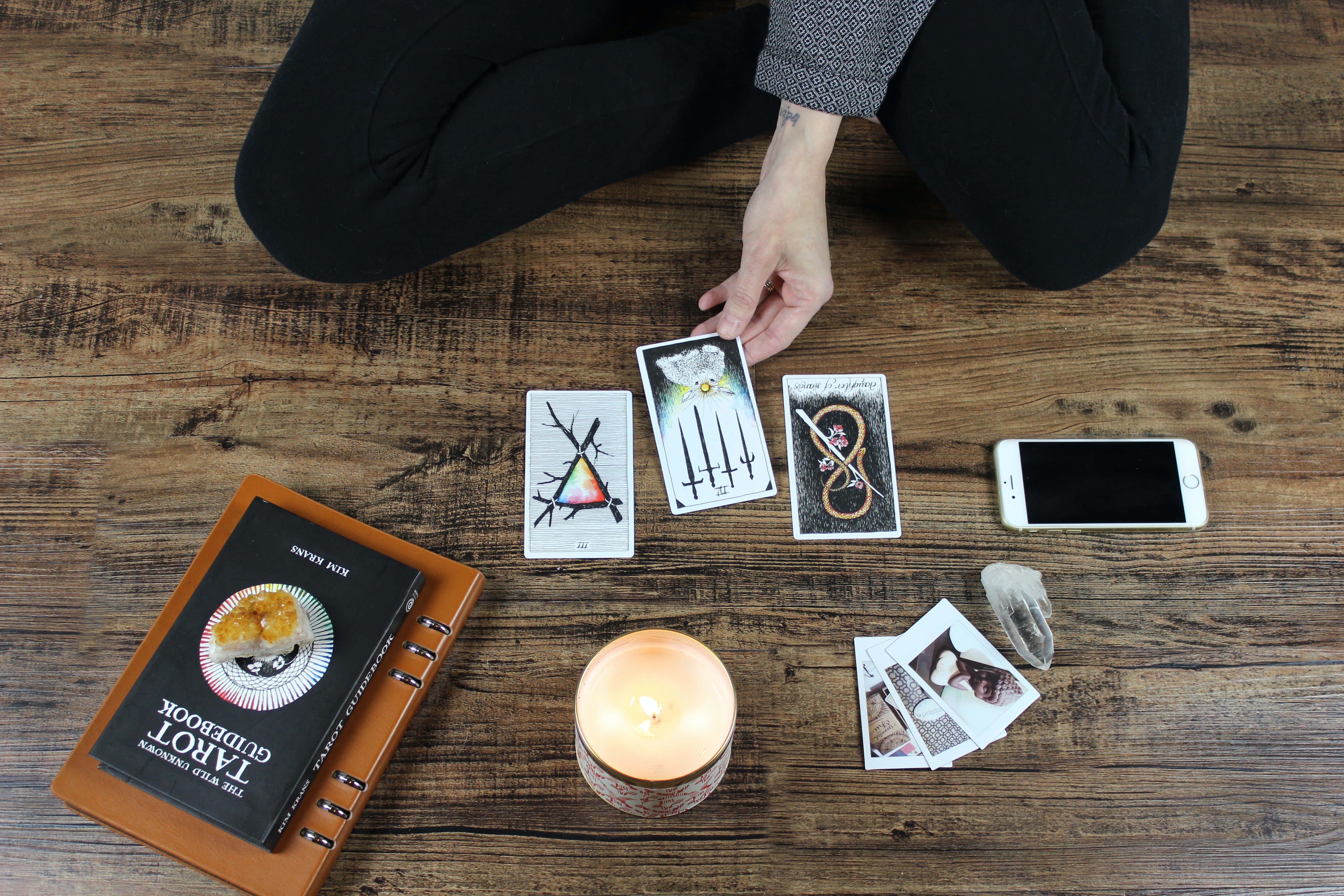 Tarot Card 桌布 壁紙 — 一個穿黑色褲子的人跪在質樸的木地板上，在點燃的蠟燭、塔羅指南書和智慧型手機旁邊排列塔羅牌。