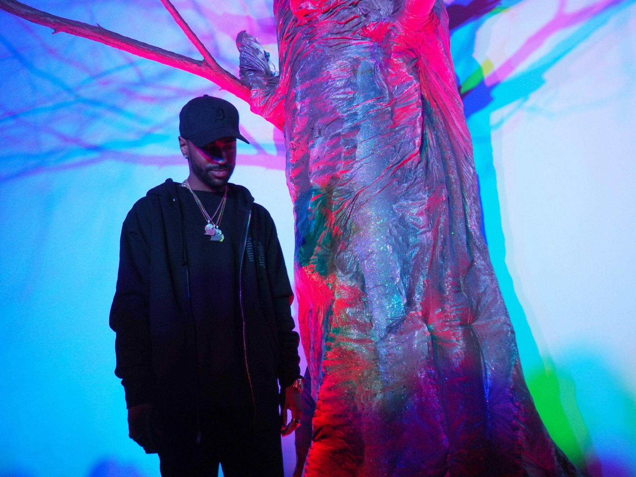Big Sean 桌布 壁紙 — 一個穿黑衣的男人站在一棵樹幹旁，樹幹在超現實的室內環境中沐浴在充滿活力的粉紅色和藍色霓虹燈光中。