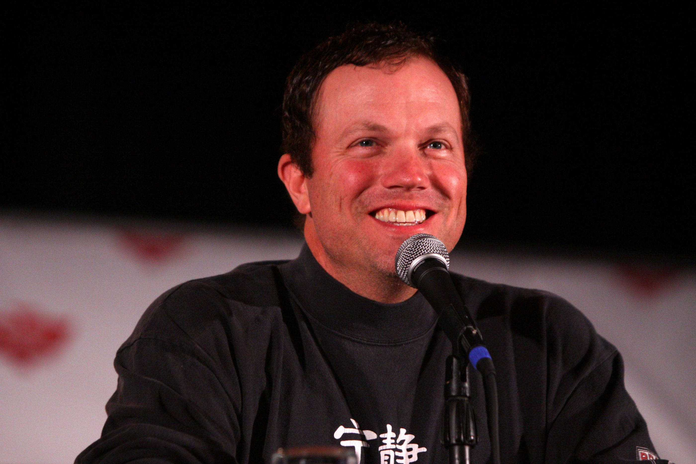 adam baldwin 桌布 壁紙 — 一個笑容燦爛的男人對著麥克風說話，背景是深色並有紅色心形圖案。
