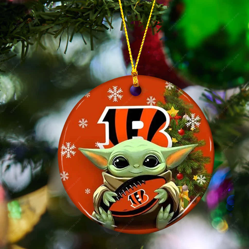 Christmas Ornaments 桌布 壁紙 — 一個節日圓形裝飾，上面有 Baby Yoda 拿著辛辛那提孟加拉虎隊的橄欖球，背景是雪白的橙色和聖誕樹枝。