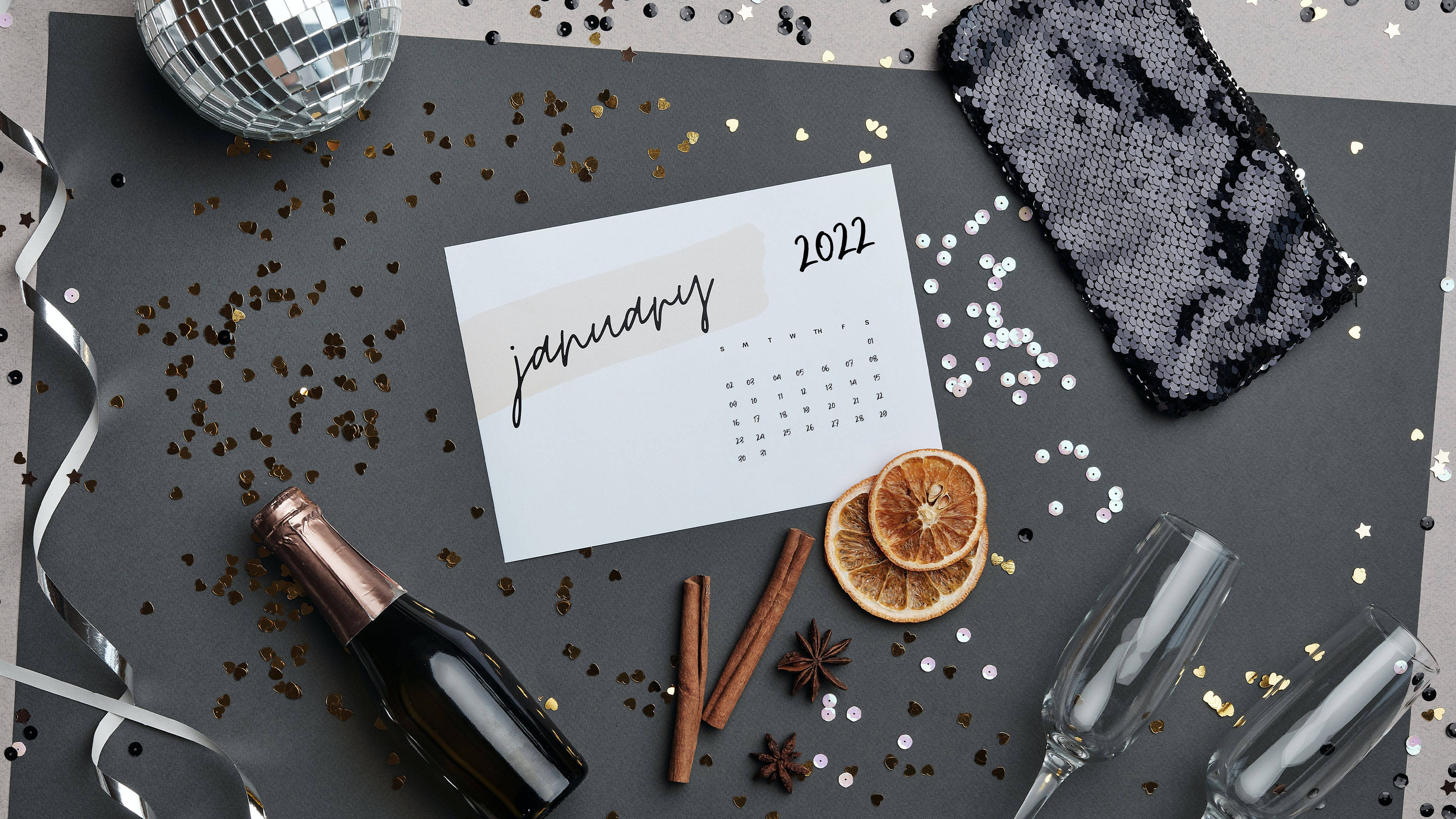 Calendar 桌布 壁紙 — 一個節日平鋪構圖，包含2022年1月日曆、香檳瓶、乾柑橘、肉桂和一個迪斯可球，放置在深色表面上。