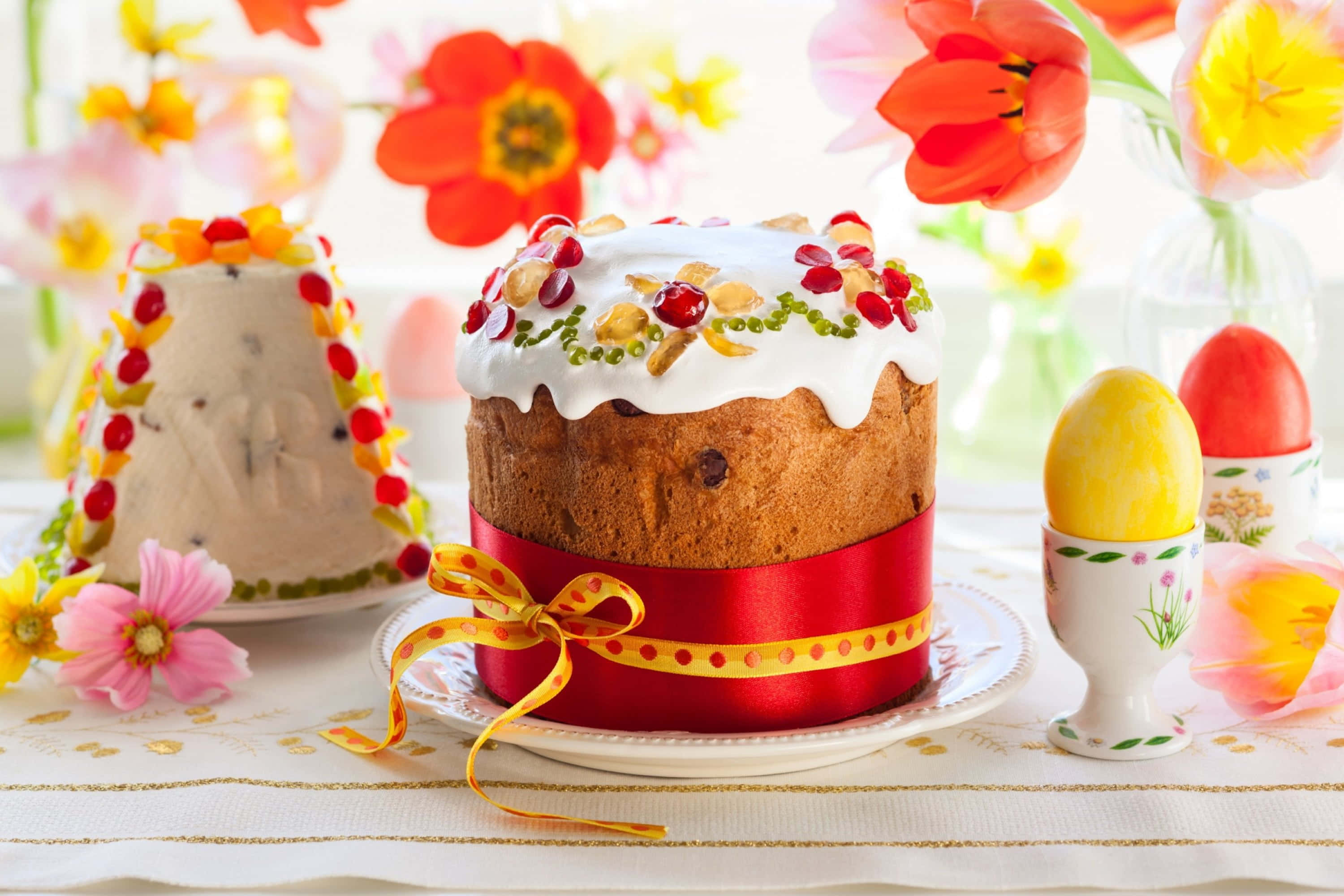Spring Desserts 桌布 壁紙 — 一個節日復活節蛋糕，裝飾著紅色漿果和綠色珠子，放在紅色緞帶上，附近有彩色雞蛋和春天花朵。