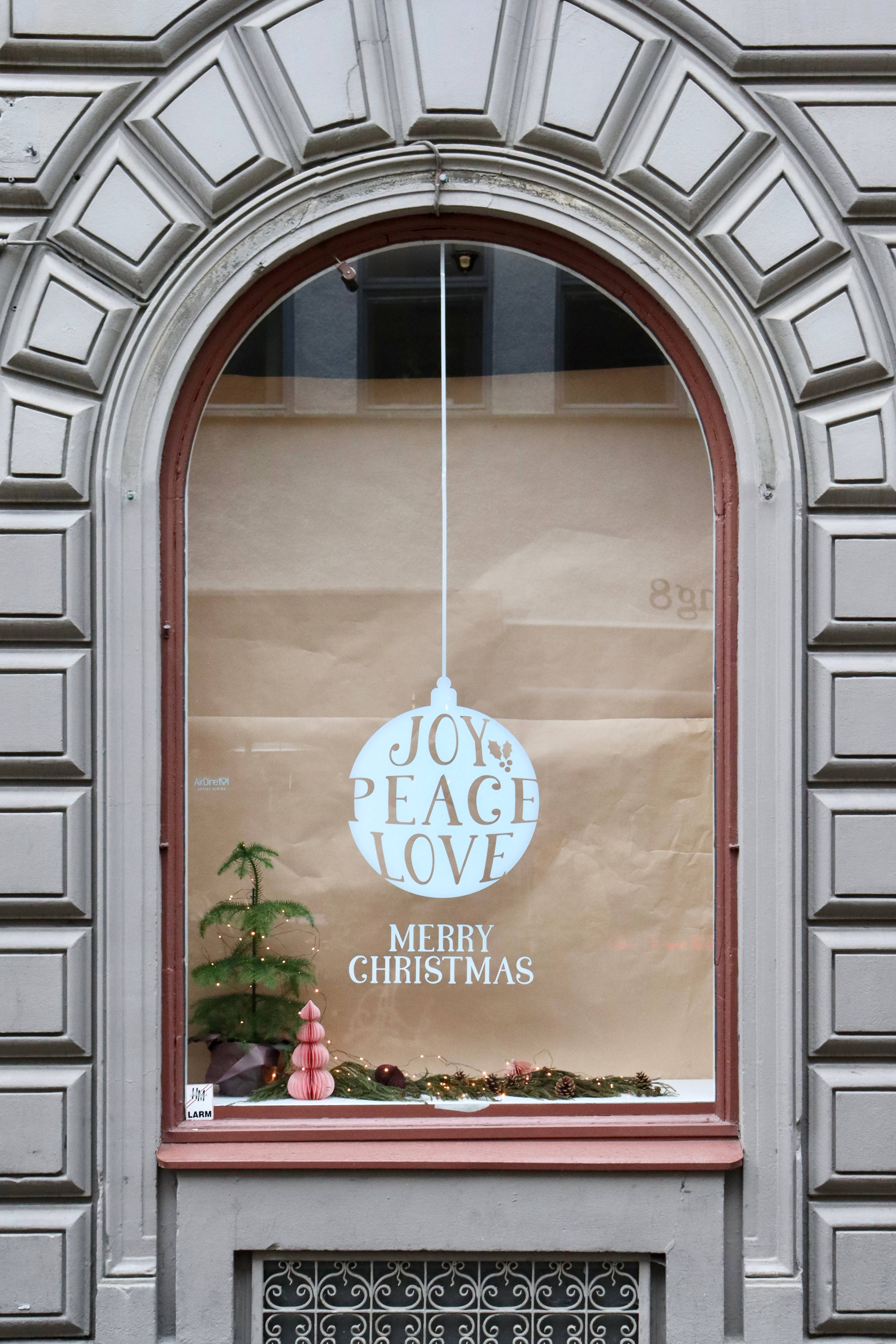聖誕快樂 桌布 壁紙 — 一個節日拱形櫥窗展示，掛有寫著 joy peace love 的裝飾品，還有一棵小聖誕樹，背景是棕色紙張。
