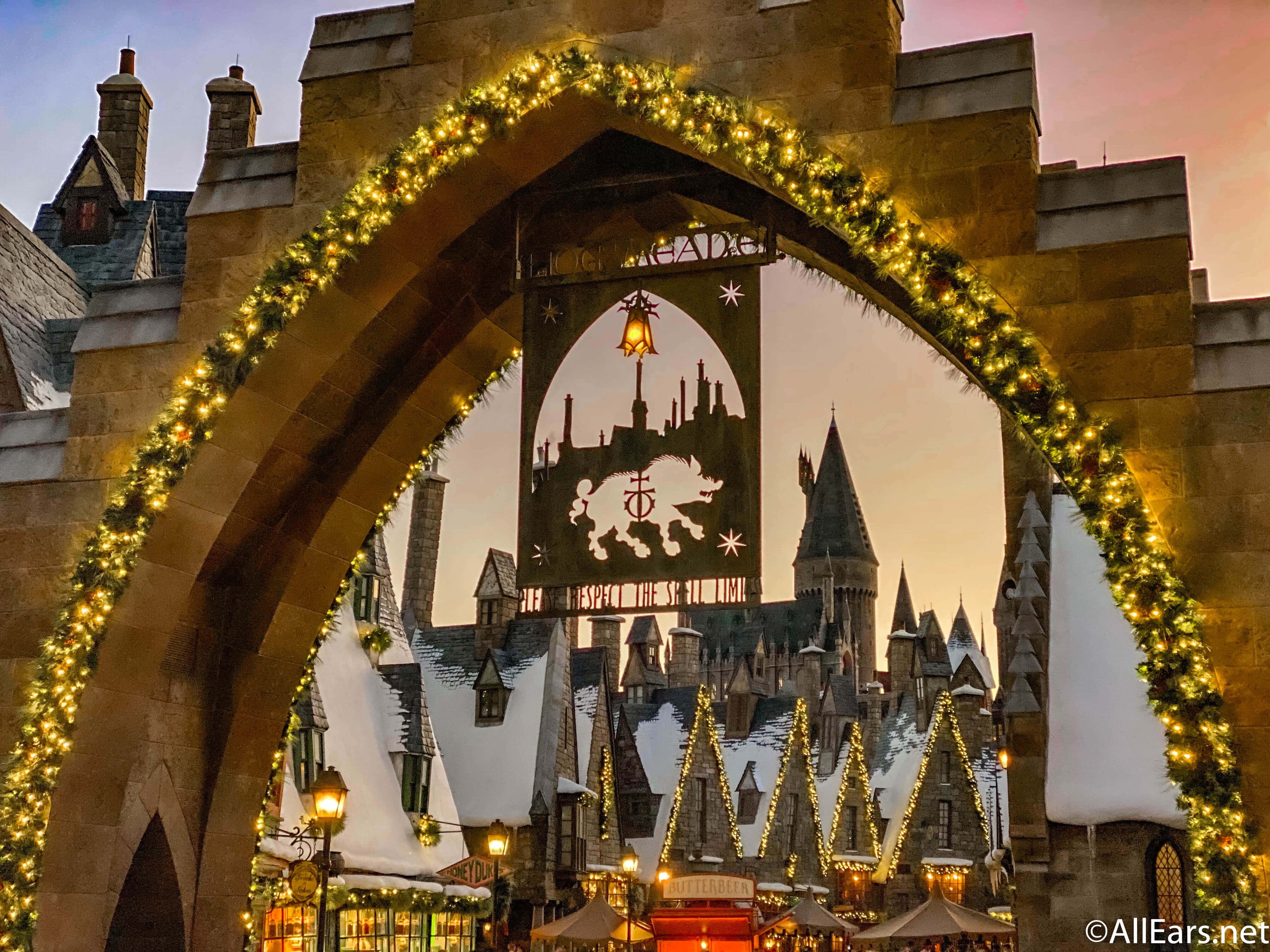 Christmas At Hogwarts 桌布 壁紙 — 一個節日拱門裝飾著發光的燈光，框住了暮色中白雪覆蓋的霍格華茲城堡。