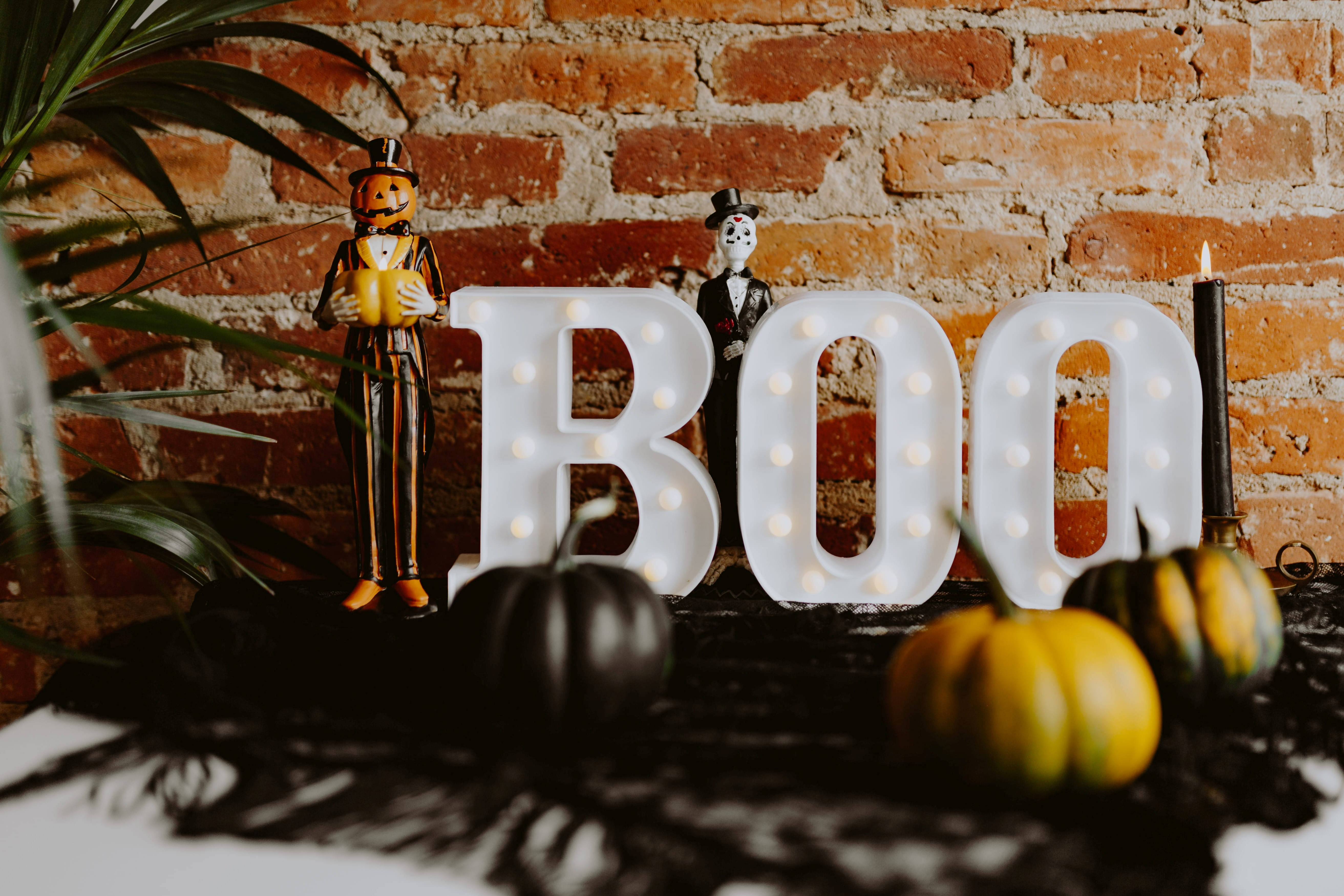 Cute Halloween 桌布 壁紙 — 一個節日氣氛濃厚的萬聖節佈置，發光的「BOO」字母、南瓜小雕像和蠟燭，背景是質樸的磚牆。