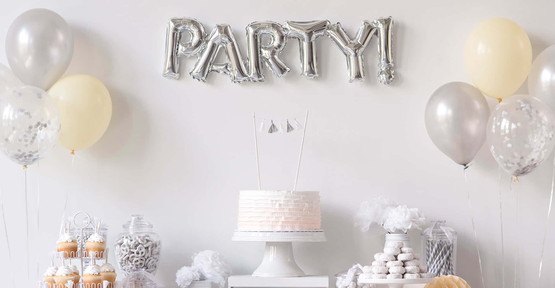 Party 桌布 壁紙 — 一個節日派對佈置，包括銀色 'PARTY!' 氣球橫幅、粉彩紙杯蛋糕和香檳色氣球，背景為白色牆壁。