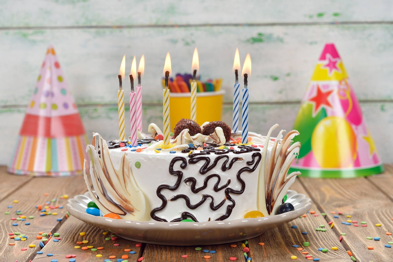 Cake 桌布 壁紙 — 一個節日生日蛋糕，上面有燃燒的蠟燭、巧克力醬和彩色糖粒，放在一個木桌上，周圍有派對帽和五彩紙屑。