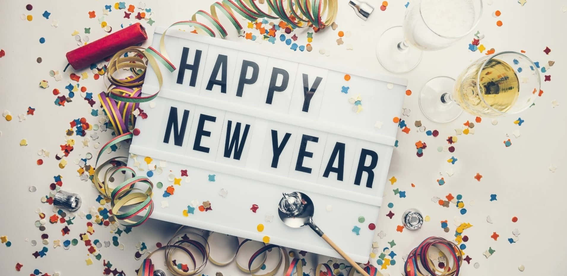 新年 桌布 壁紙 — 一個節日白色燈箱，顯示「HAPPY NEW YEAR」，周圍環繞著五彩紙屑、絲帶、香檳杯和開瓶器，位於明亮的表面上。