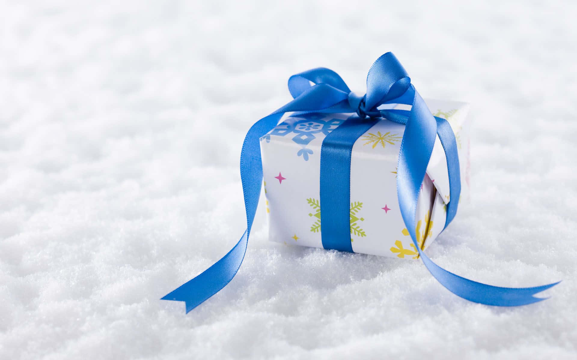 Gift 桌布 壁紙 — 一個節日禮物盒，用白色雪花圖案紙包裝，並繫上亮藍色絲帶，放在新雪上。