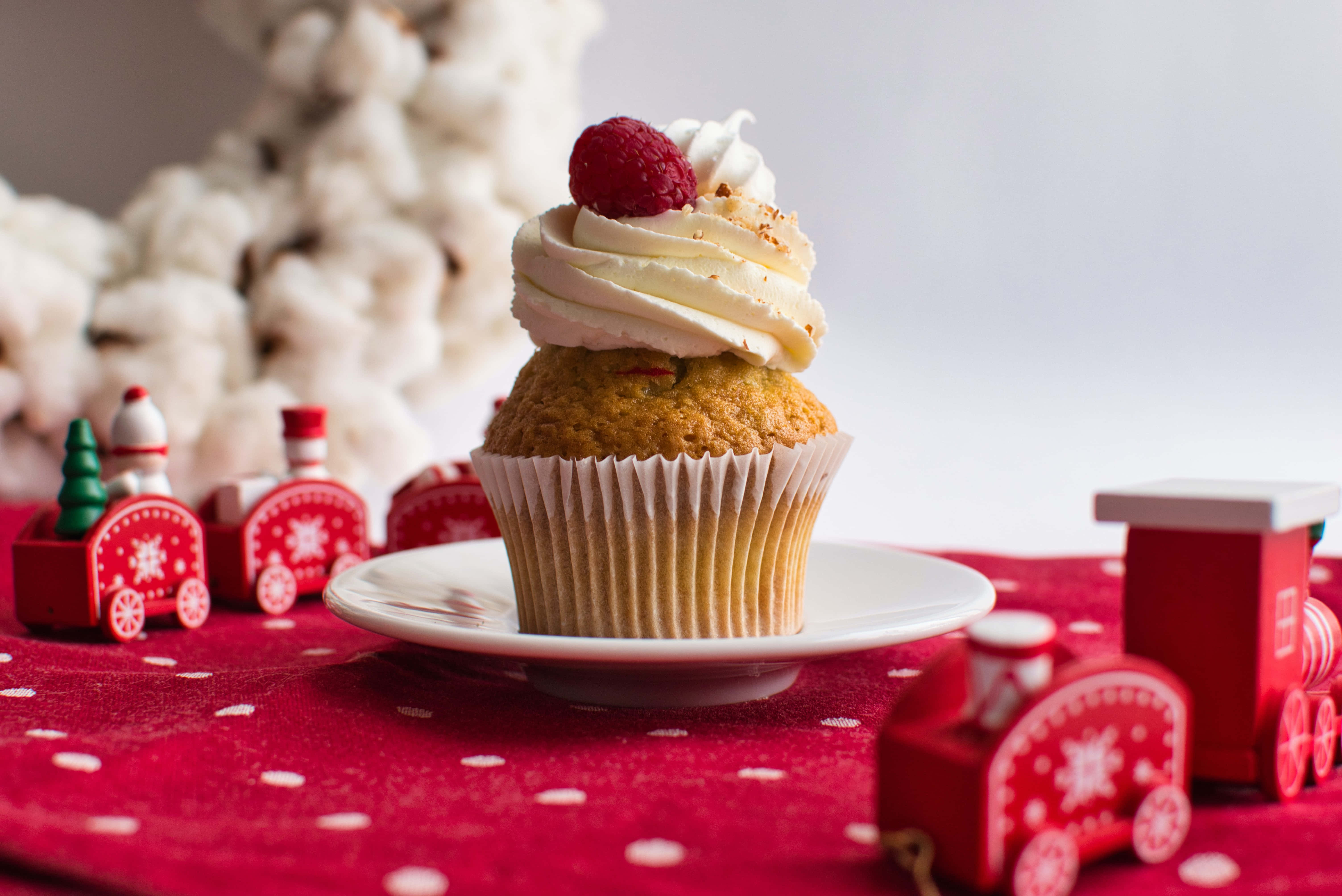 Cupcake 桌布 壁紙 — 一個節日紙杯蛋糕，上面有奶油糖霜和覆盆子，放在白色盤子上，置於紅色圓點布上，周圍環繞著迷你紅色玩具火車和棉花樹裝飾。