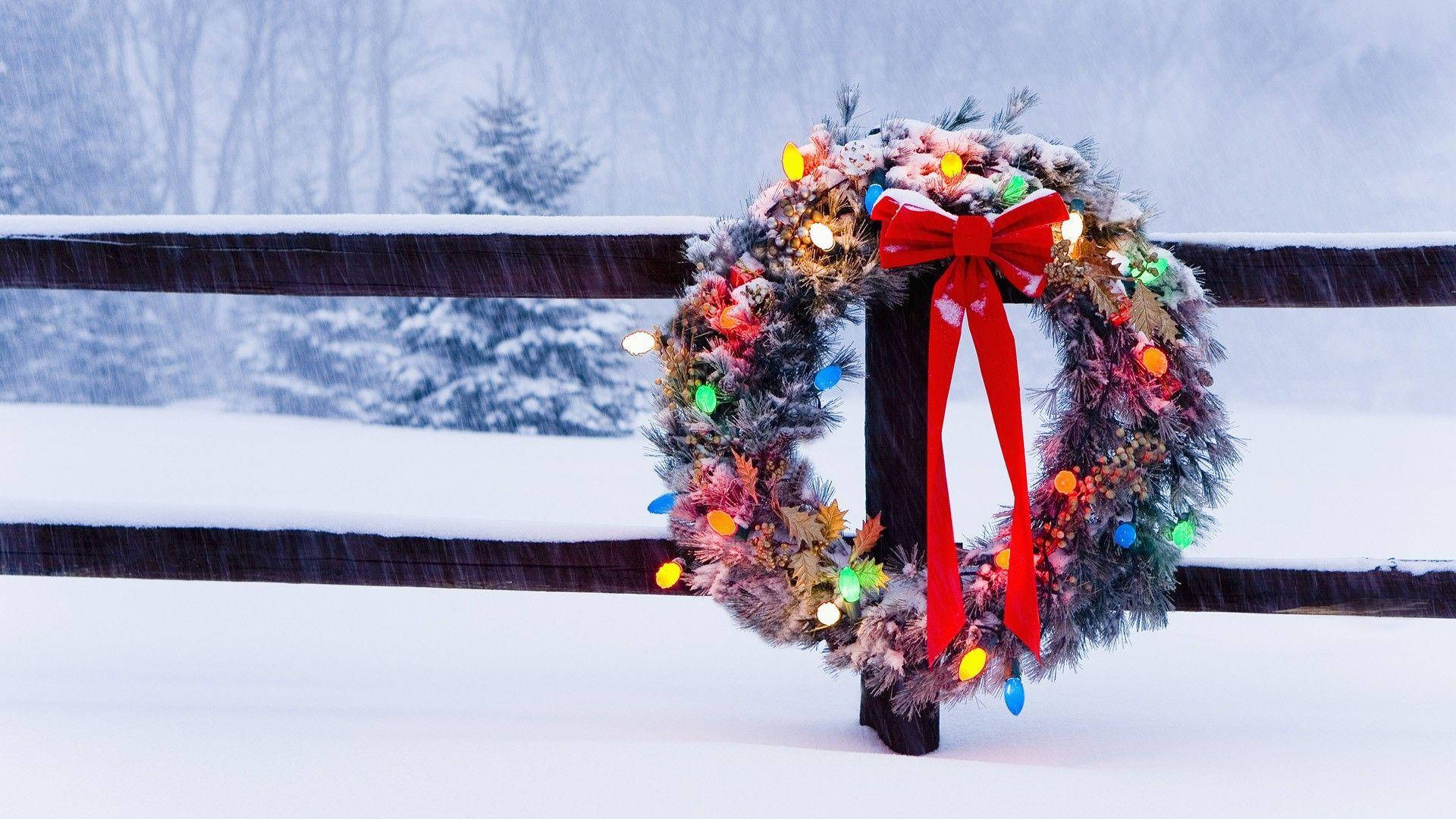 Wreath 桌布 壁紙 — 一個節日聖誕花環，帶有閃爍的燈光和紅色蝴蝶結，掛在冬季景觀中積雪覆蓋的木柵欄上。