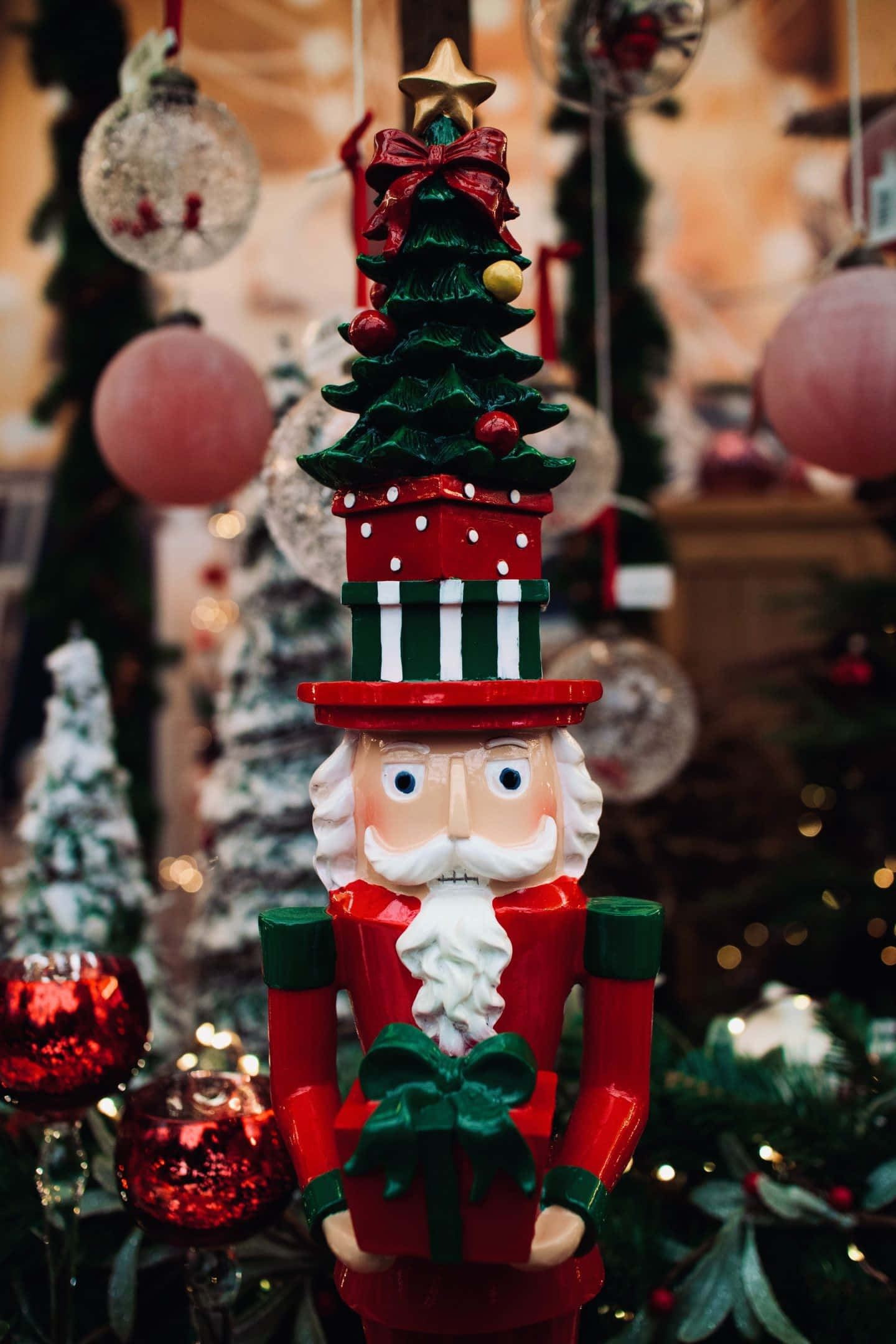 Red Christmas iPhone 桌布 壁紙 — 一個節日胡桃夾子士兵手持一個禮物盒，上面放著迷你聖誕樹和紅色蝴蝶結，背景是模糊的節日裝飾。