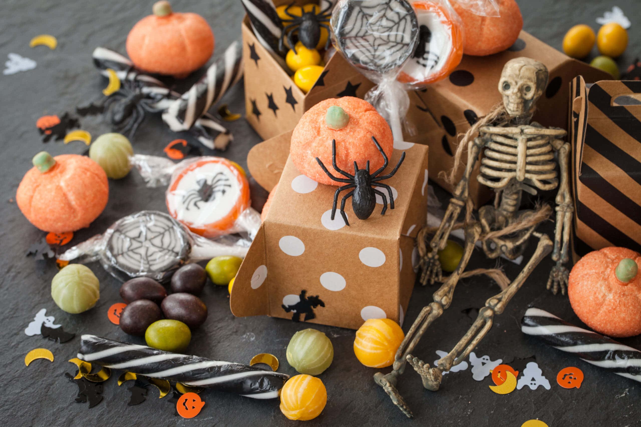 Halloween Treats 桌布 壁紙 — 一個節日般的萬聖節擺設，包括糖果南瓜、蜘蛛、骷髏和條紋零食，散落在深色石板表面上。