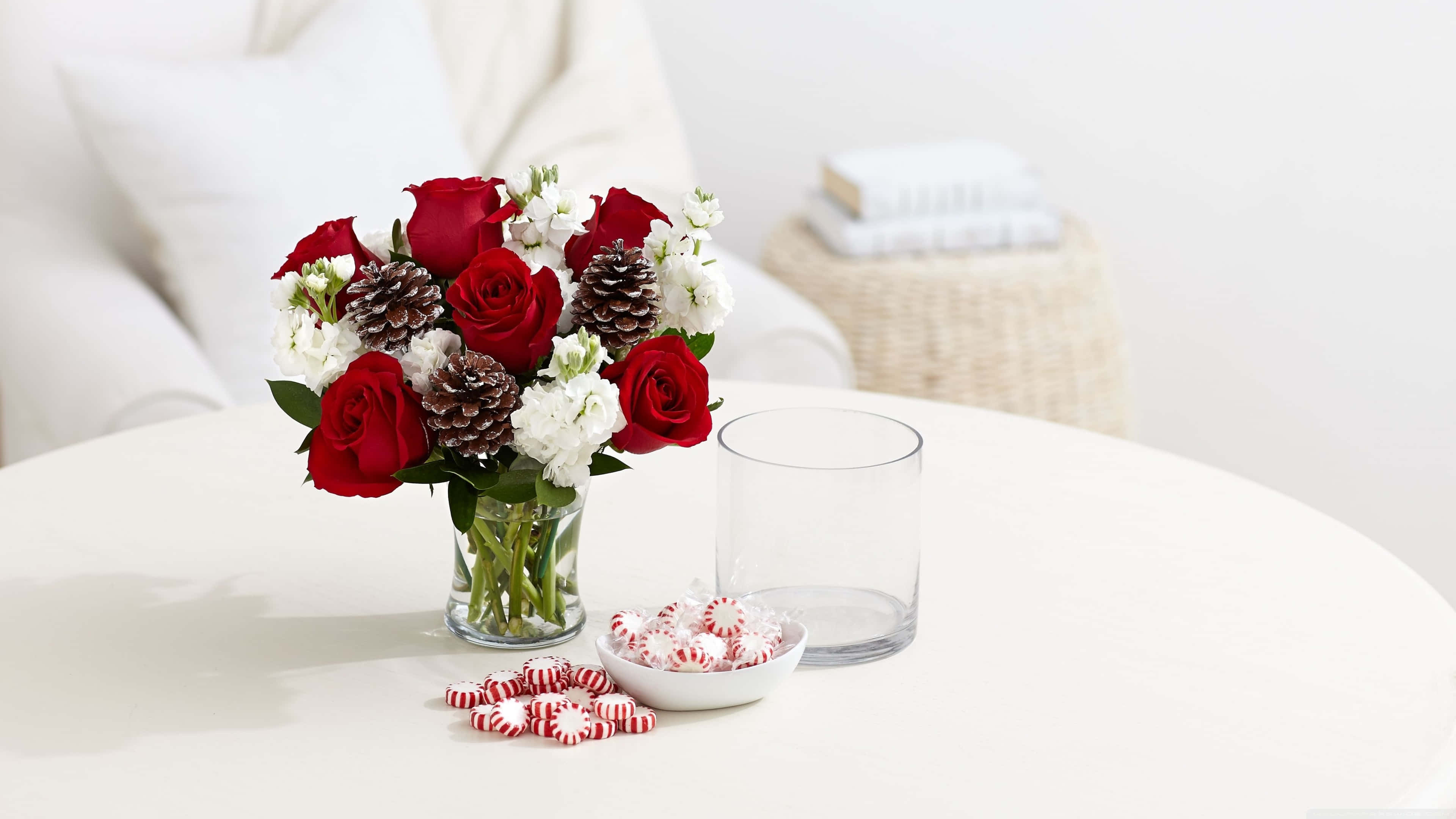Red And White Roses 桌布 壁紙 — 一個節日花束，包含紅玫瑰、白花和松果，放在玻璃花瓶裡，置於白色桌子上，旁邊有薄荷糖和一個透明玻璃杯。