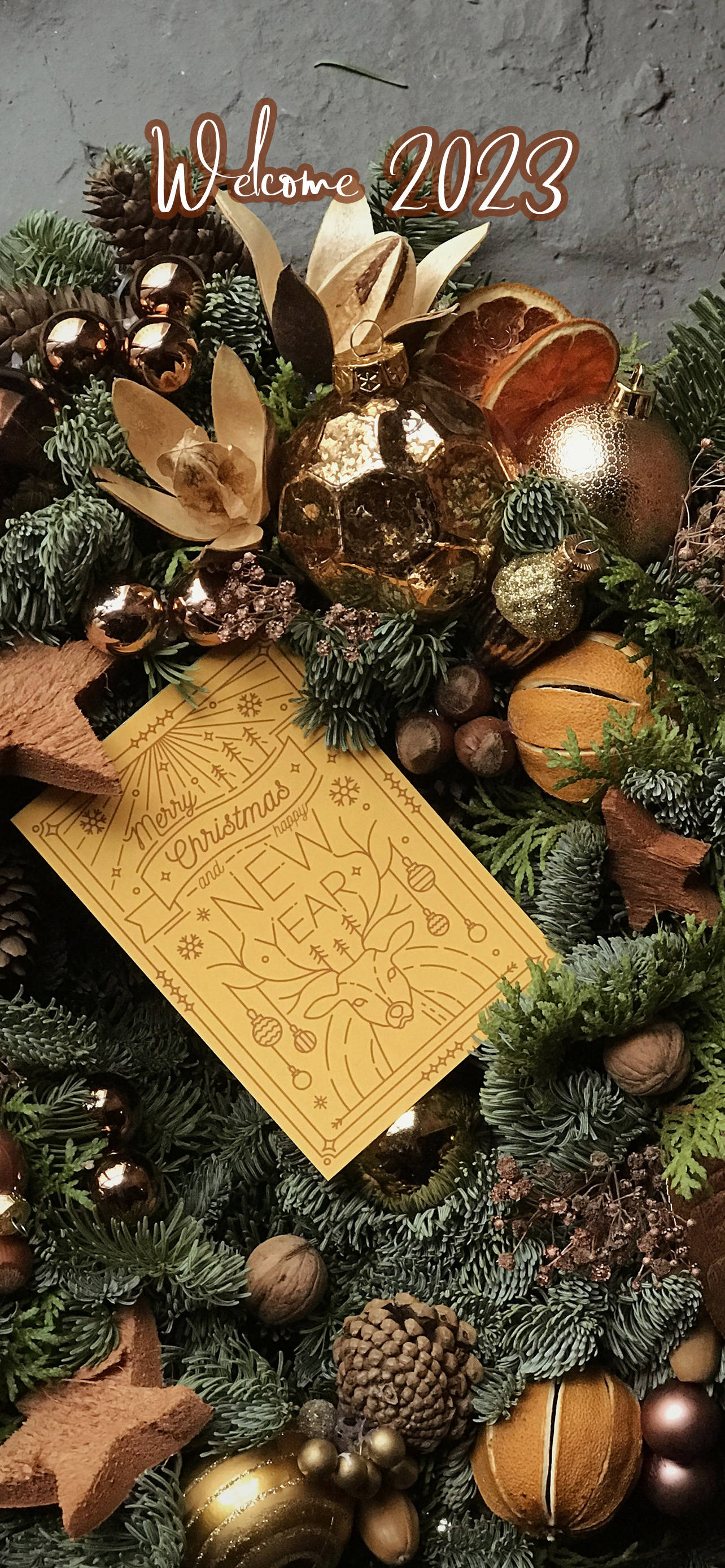 Greetings 桌布 壁紙 — 一個節日花環，裝飾著松果、金色飾品、乾柑橘片和一張金色卡片，上面寫著「歡迎2023」，背景是紋理灰色牆壁。