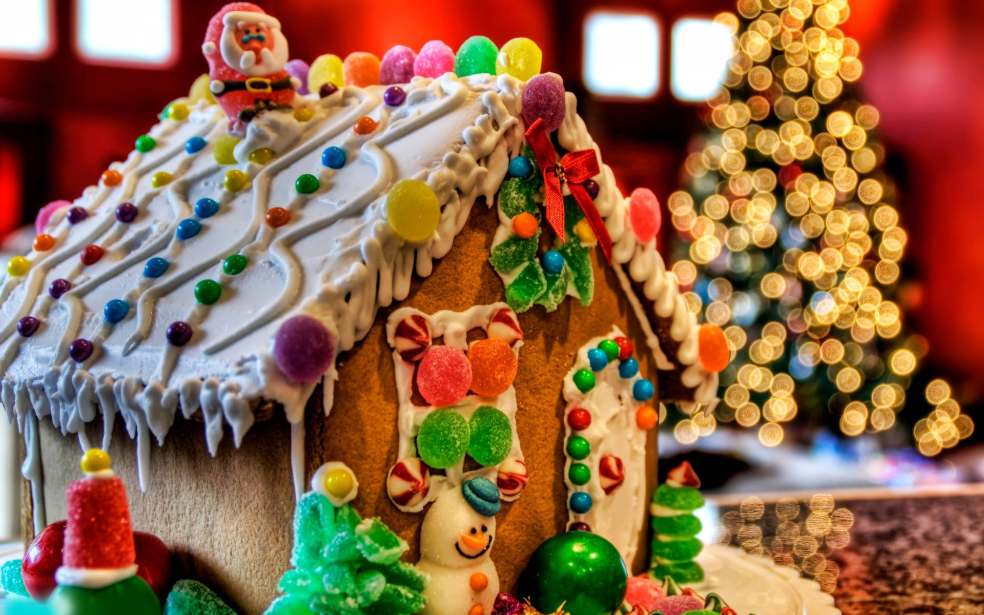 Gingerbread House 桌布 壁紙 — 一個節日薑餅屋，裝飾著色彩繽紛的糖果和聖誕老人公仔，背景是模糊的聖誕樹和閃爍的燈光。