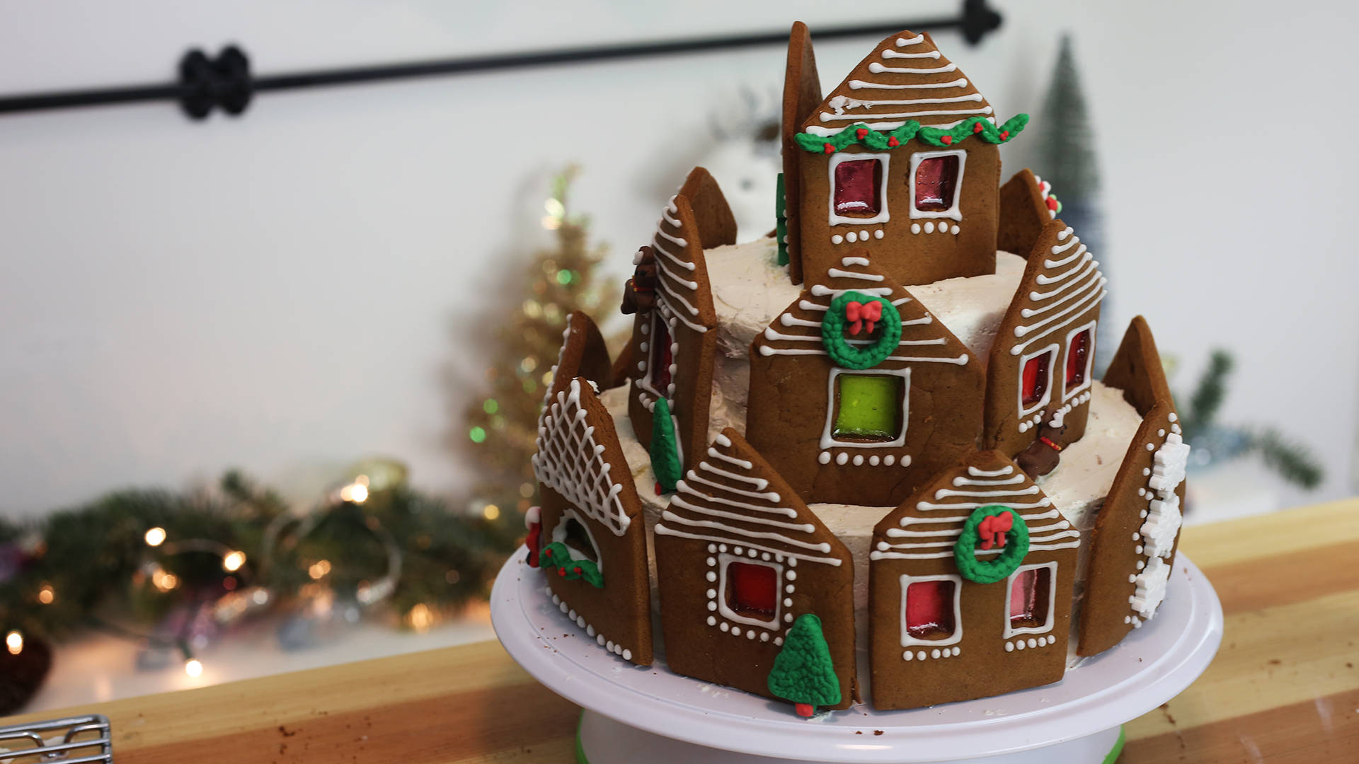 Gingerbread House 桌布 壁紙 — 一個節日薑餅屋城堡，飾有糖霜細節和糖果窗戶，放在白色盤子上，背景是柔和燈光的假日場景。
