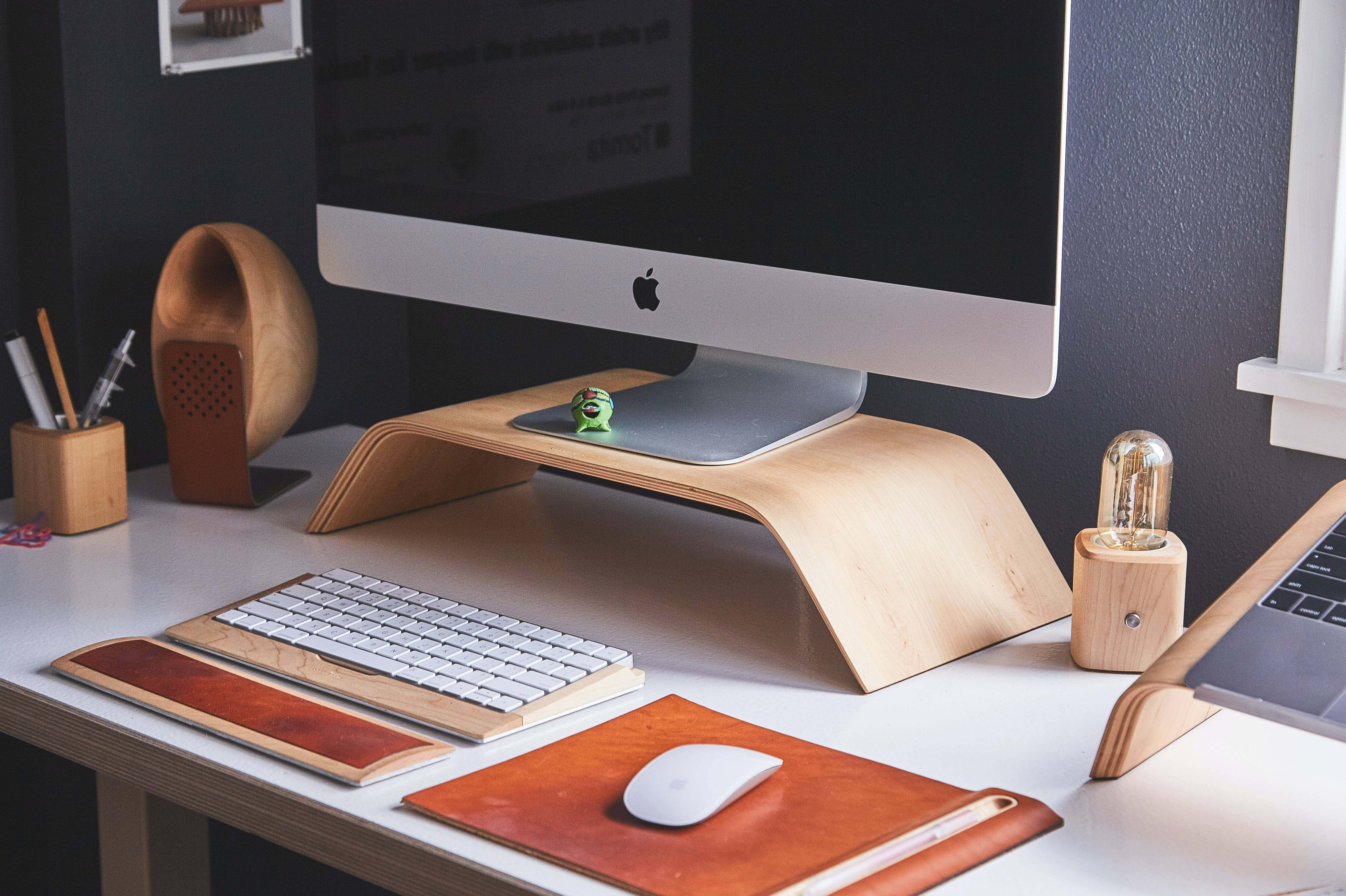 Mac 桌布 壁紙 — 一個簡約現代的辦公桌設置，配有木製支架，放置 iMac 鍵盤、滑鼠和溫暖的皮革配件，在柔和環境光下。