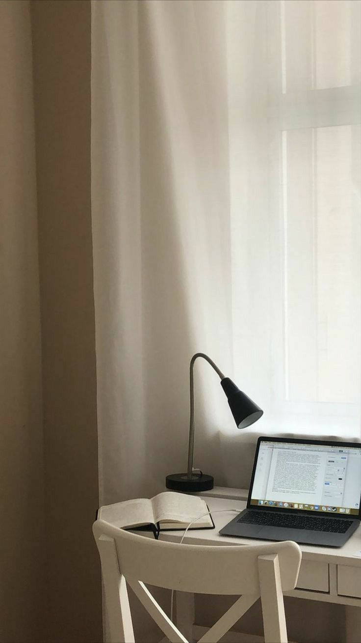 MacBook 動態圖片 桌布 壁紙 — 一個簡約的工作空間，白色桌子上有一部打開的筆記型電腦，旁邊有一本打開的書和一個黑色檯燈，柔和自然光從薄窗簾透入。