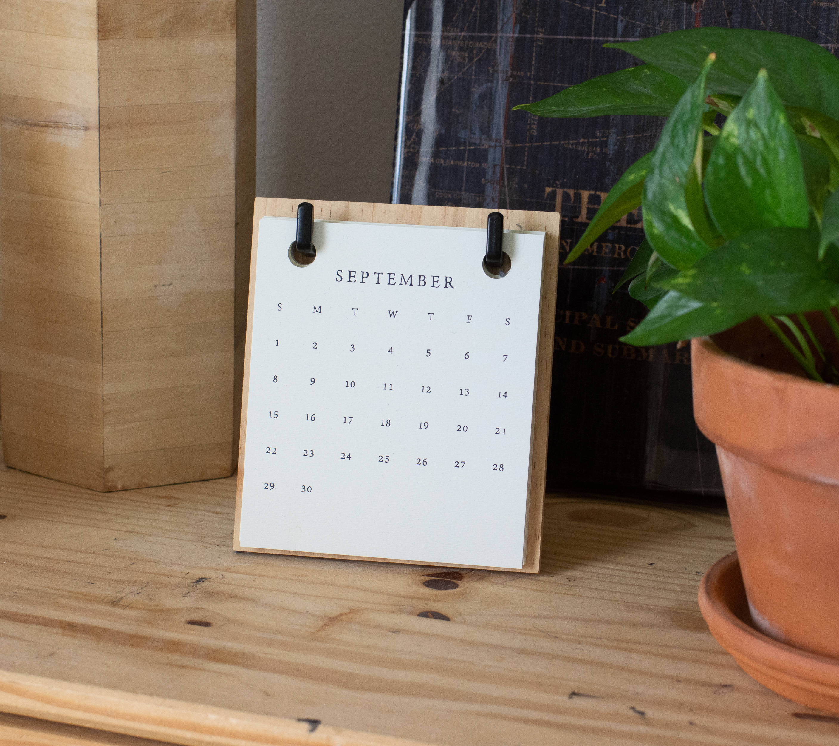 february 2021 calendar 桌布 壁紙 — 一個簡約的木製桌面日曆，顯示九月，放在淺色木架上，旁邊有一盆綠蘿。