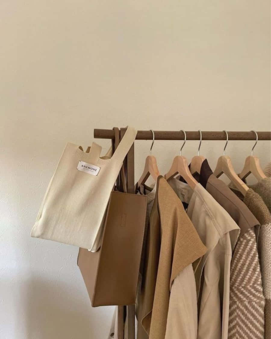 Korean Beige Aesthetic 桌布 壁紙 — 一個簡約的木製衣架掛著米色和棕色的毛衣和一個相配的手提袋，背景是素牆。