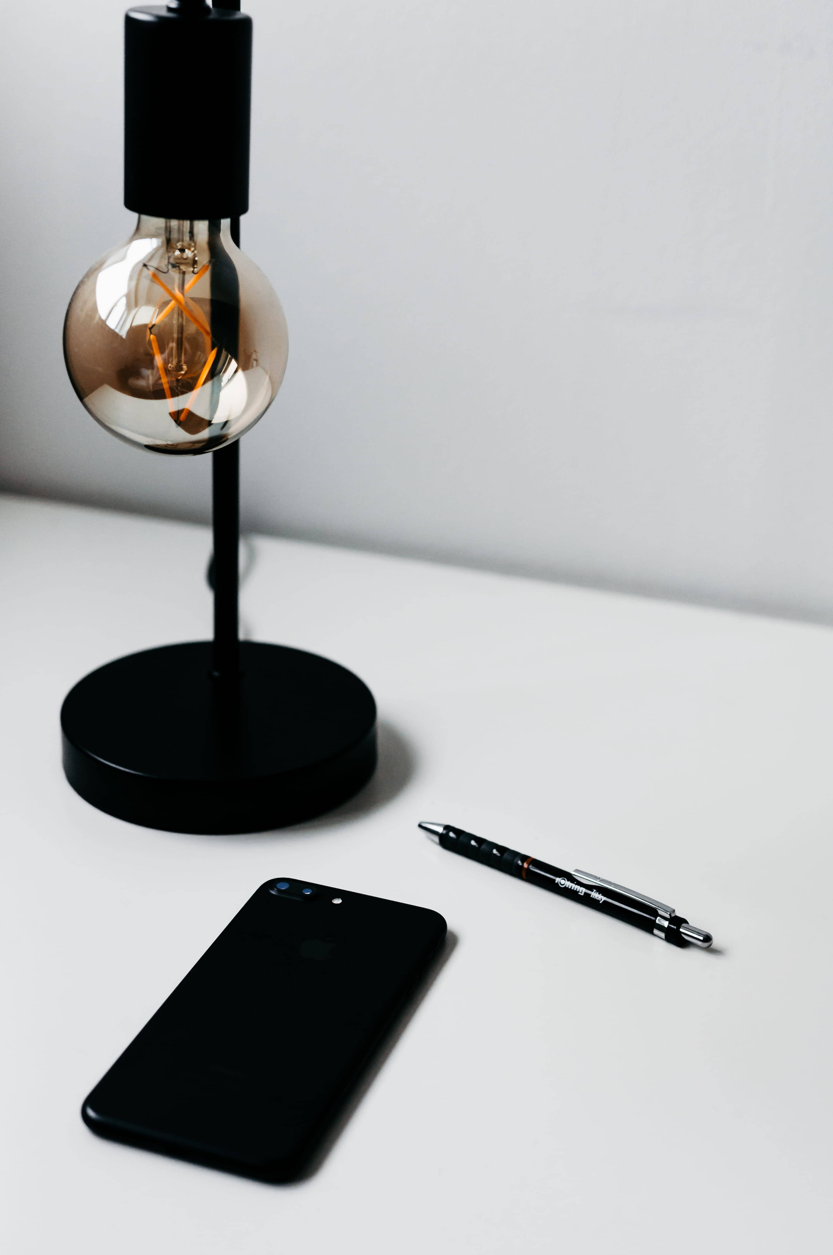 iPhone 桌布 壁紙 — 一個簡約的桌面設置，包括黑色愛迪生燈泡燈、電話和筆，放在白色表面上。
