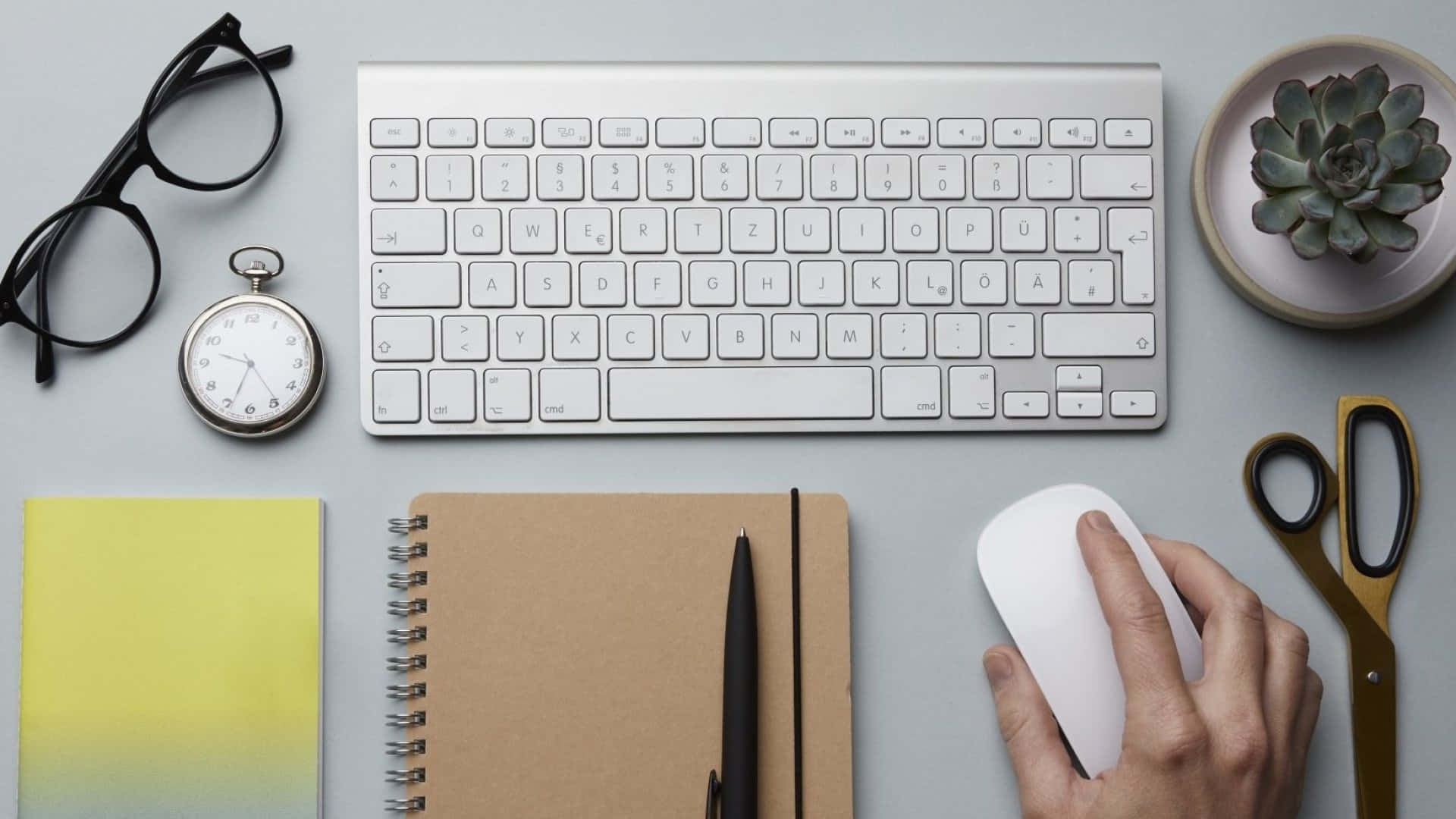 Computer Mouse 桌布 壁紙 — 一個簡約的桌面設置，有一隻手在使用白色滑鼠，旁邊有鍵盤、筆記本、眼鏡和一棵多肉植物。
